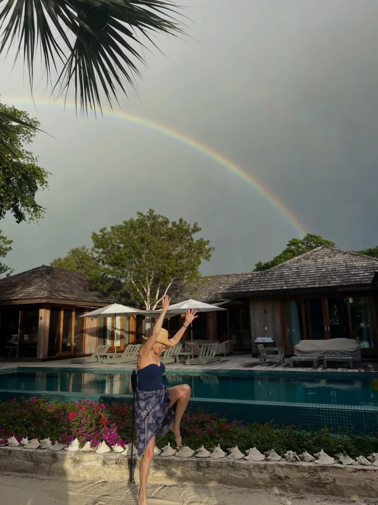 She wore a navy one-piece swimsuit paired with a lightweight blue patterned cover-up and a straw sun hat, smiling as she sat along the water’s edge.