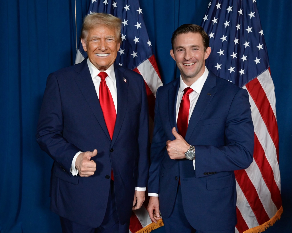 Donald Trump and a younger man in matching suits and red ties stand in front of US flags, giving thumbs up.