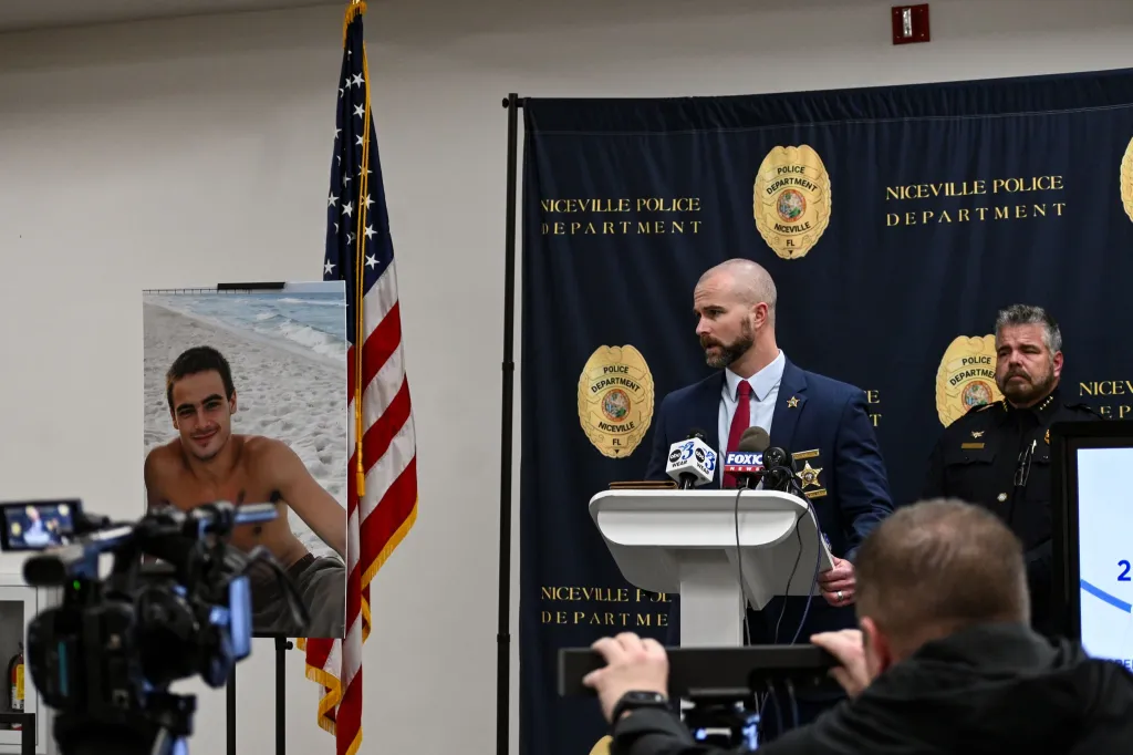 A man in a suit speaks at a podium next to an American flag, with an inset photo of a young man on a beach.