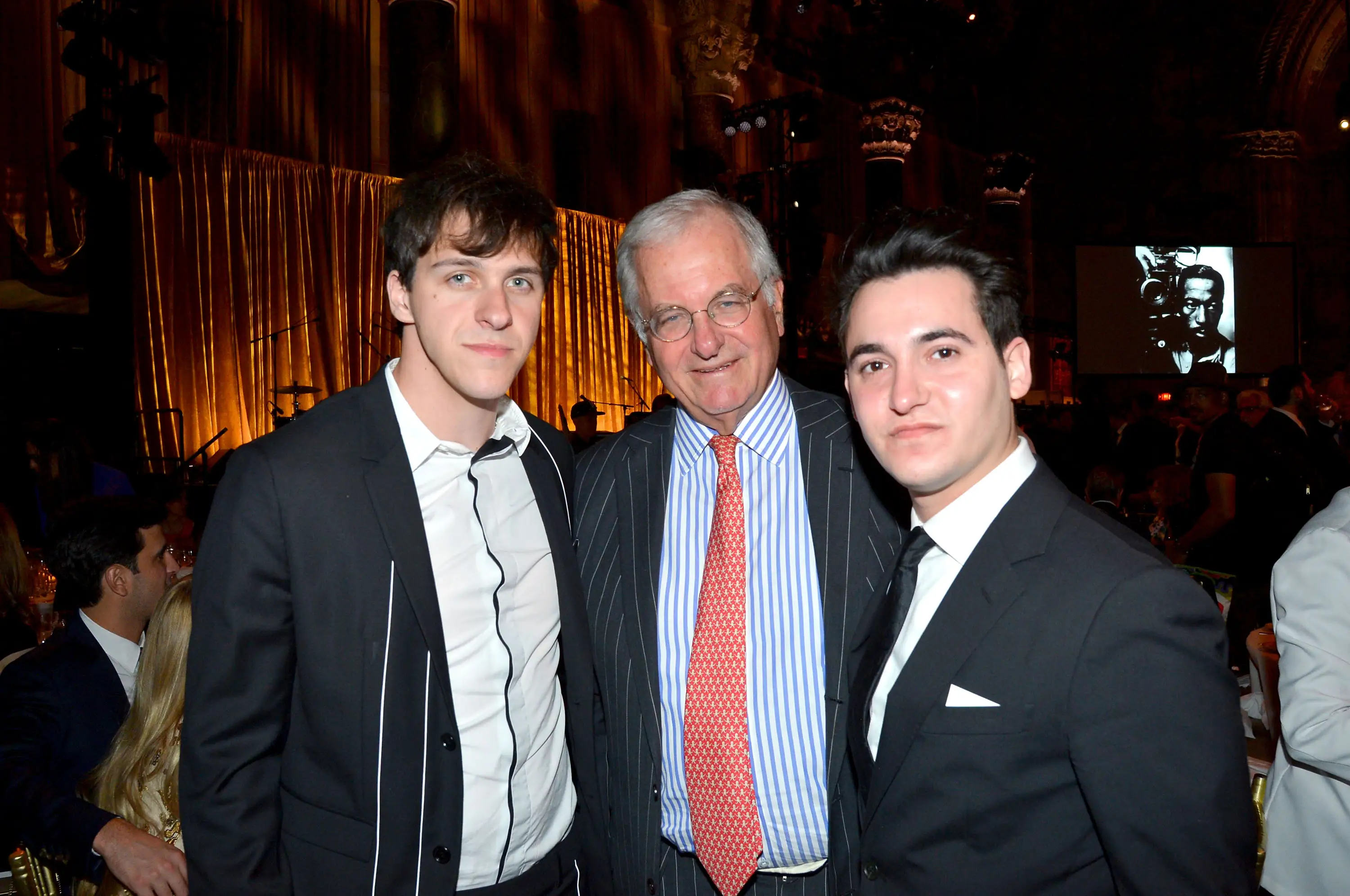 Diesel Peltz standing on the far left wearing a black jacket over a white button down, in the middle an old man is wearing a blue striped button down with an orange tie and black suit jacket, on the far right a younger man is wearing a black jacket with white button down and tie