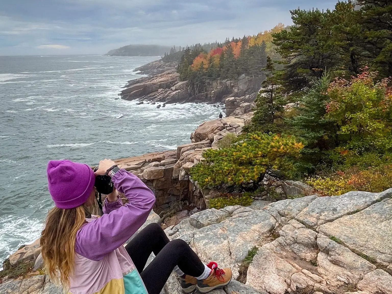 Emily sits on a rock by the water and takes photos of the fall foliage. Her backpack, covered in patches, sits next to her on the rock.