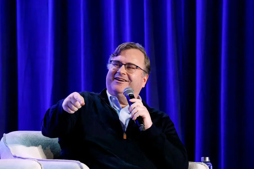 Reid Hoffman onstage speaking at an event with a black sweater, sitting and holding a microphone in front of a blue curtain.