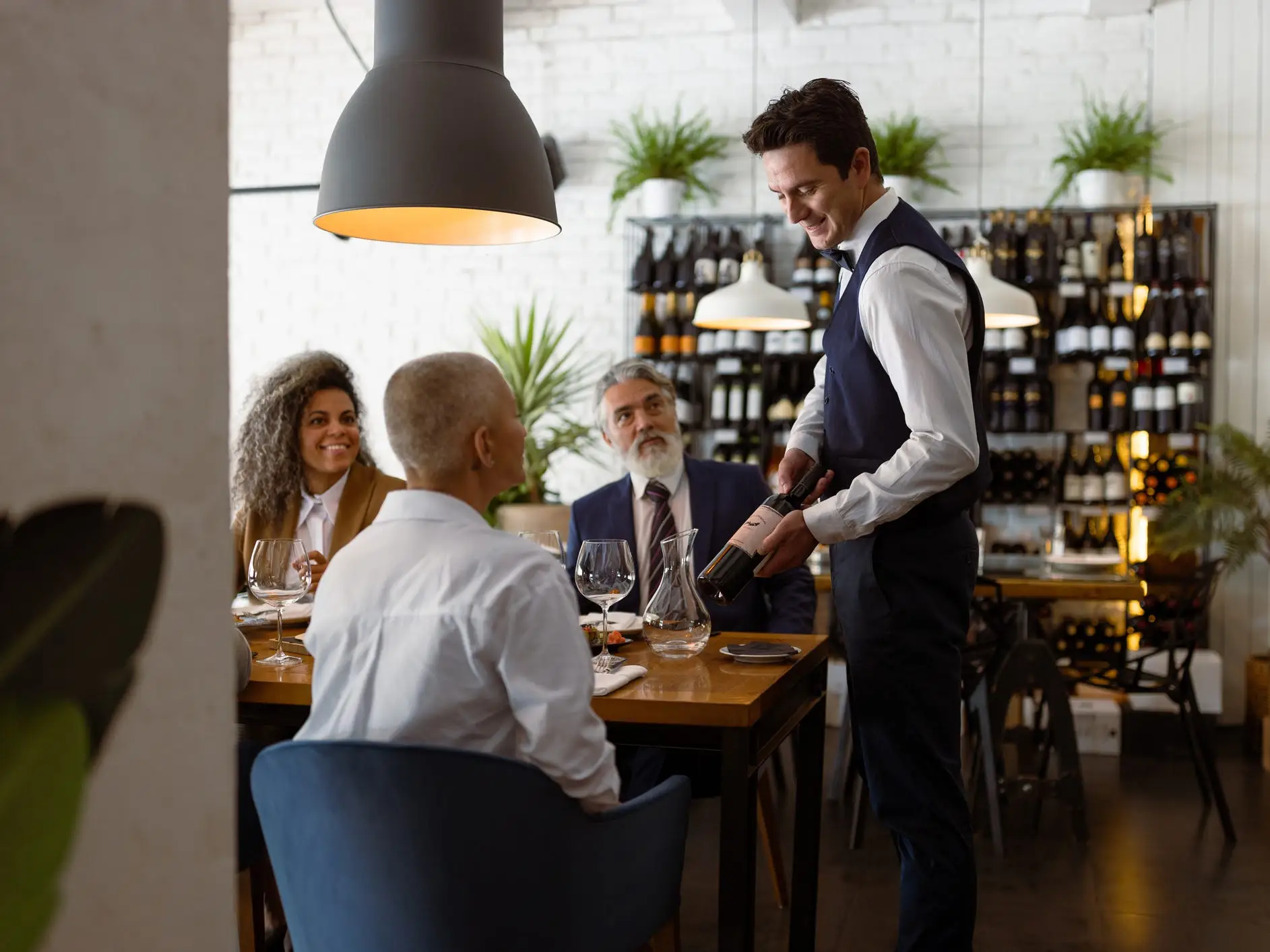 A waiter showing two men and a woman a bottle of wine as they sit at a table in a restaurant.