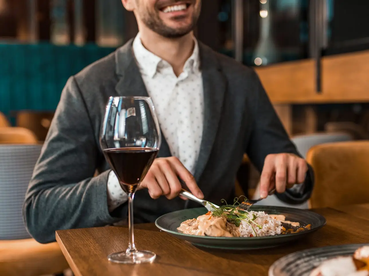 A man, dressed in a button-down jacket and a sports jacket, cuts his meal at a restaurant with a glass of red wine in front of him.
