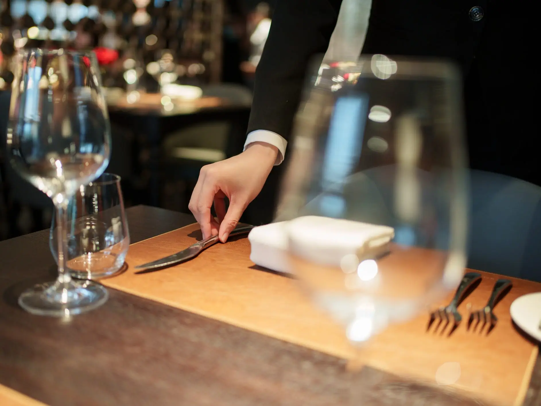 A hand places a knife on a table set with wine glasses, placemats, a napkin, and silverware at a restaurant.