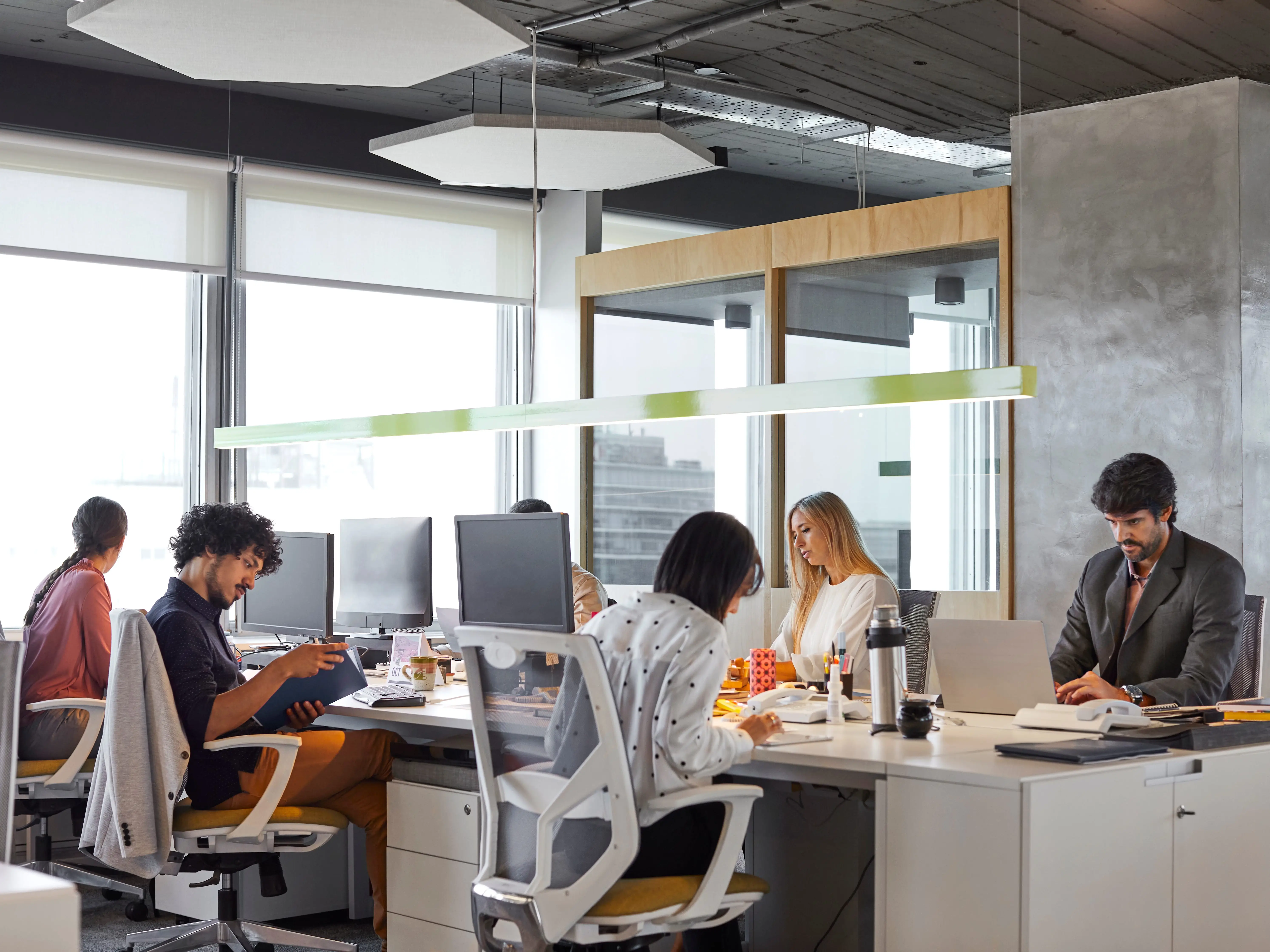 Six people sitting at a communal desk in an office doing work.