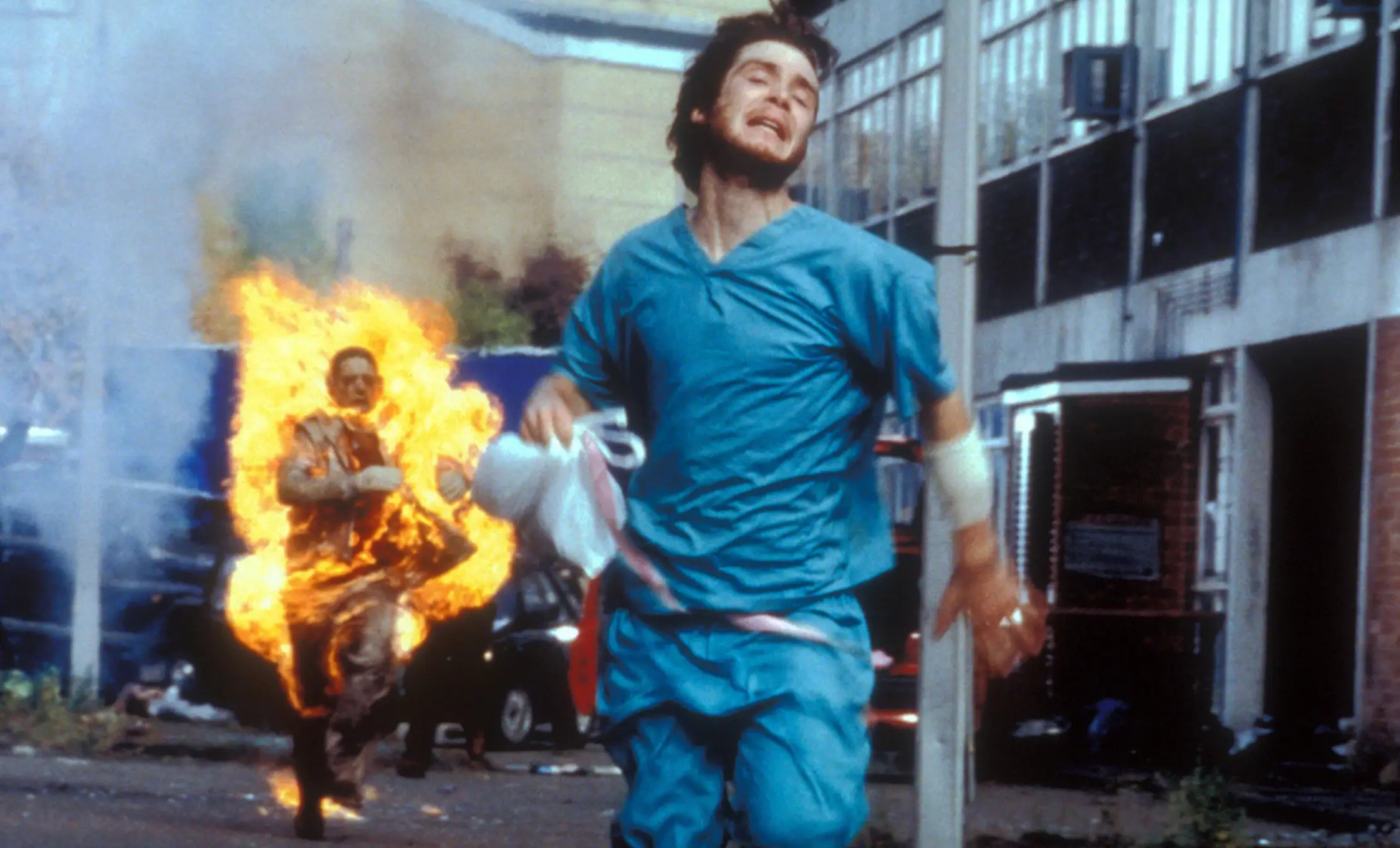 A man with long brown hair wearing blue hospital scrubs is running away from a zombie on fire down a street.
