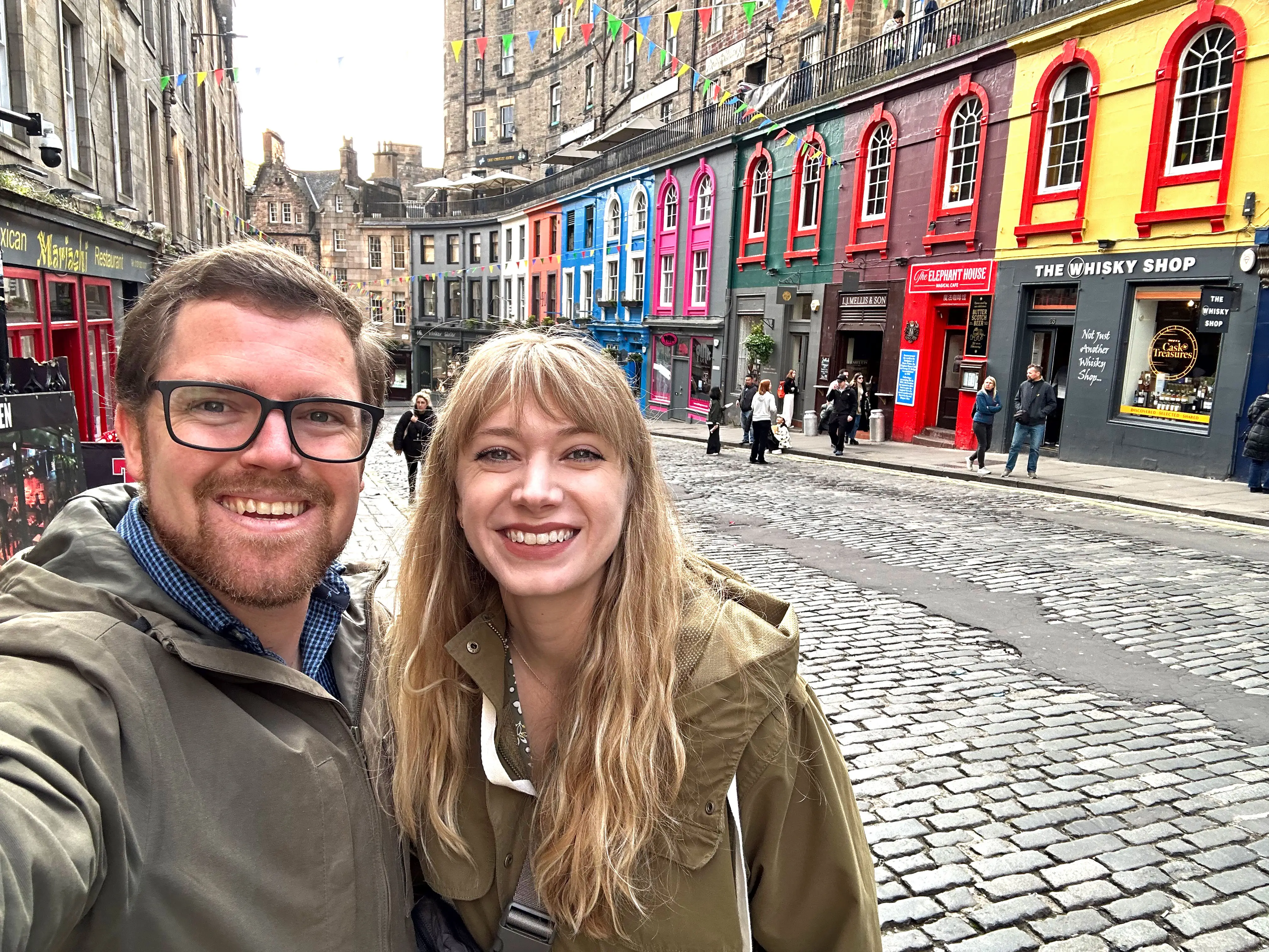 Emily and her husband take a selfie on a cobblestone street lined with colorful shops.