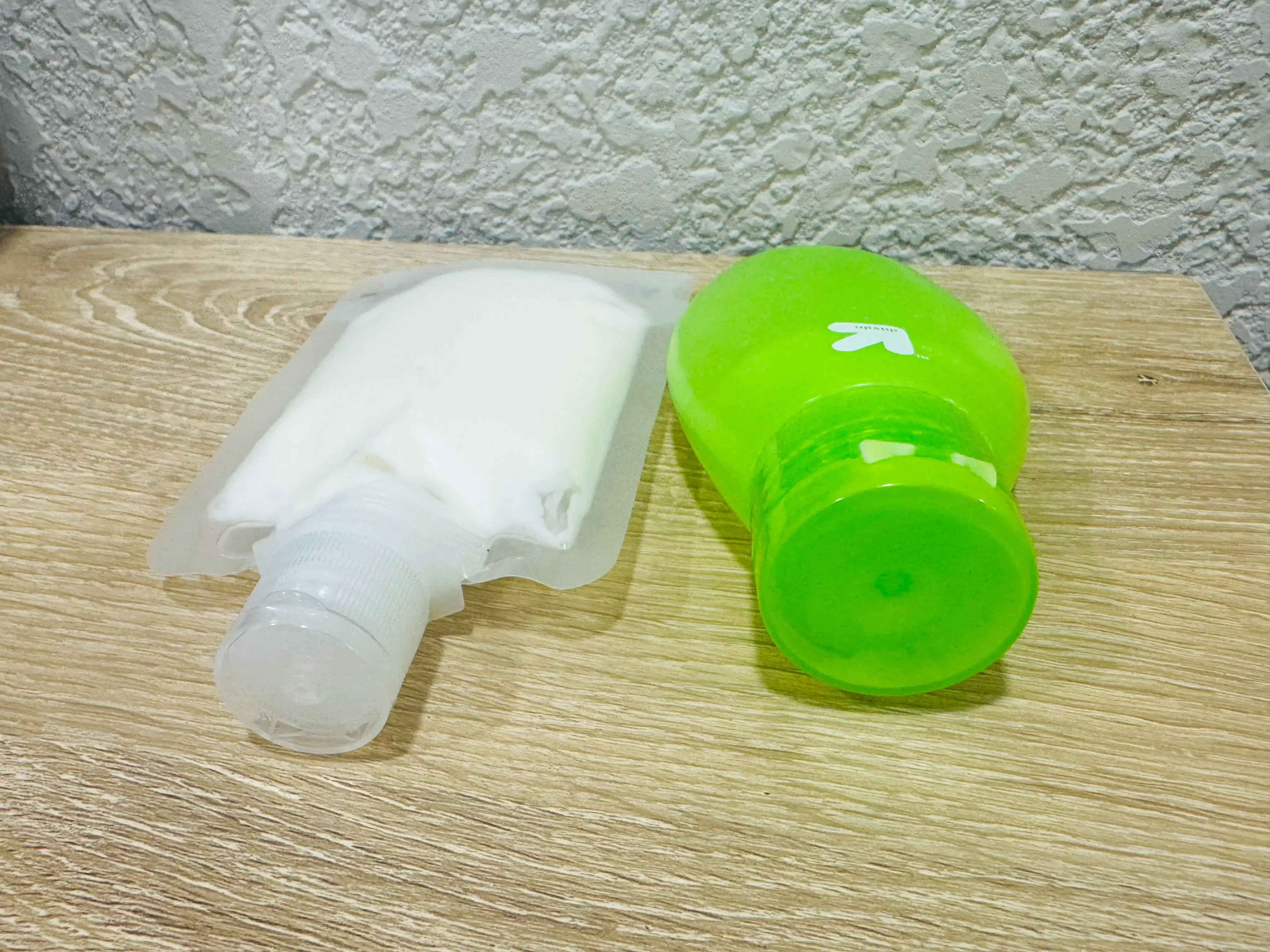 A clear, flat travel pouch with lotion inside and a more bulky, green travel container on a wooden table.