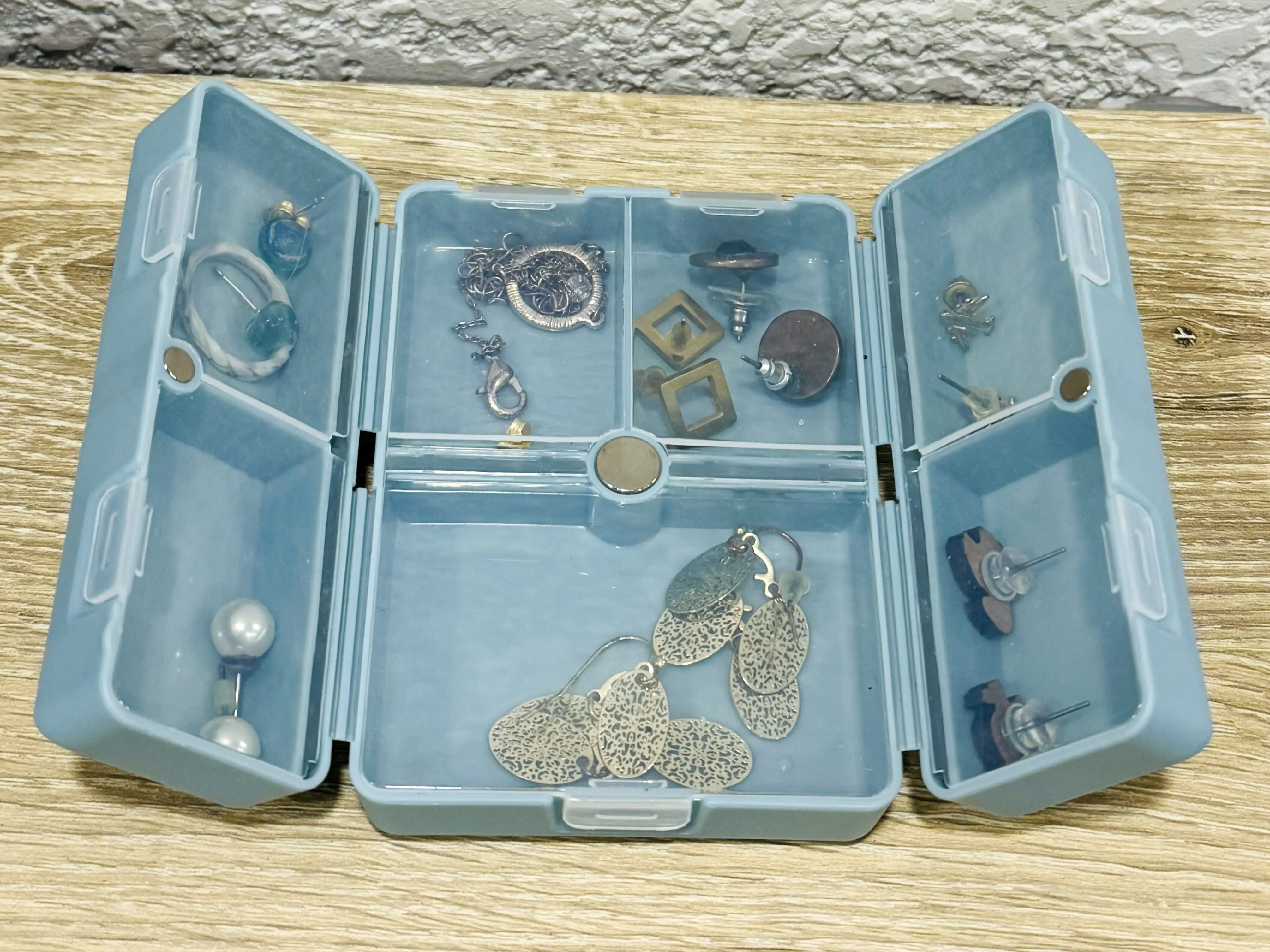 A blue plastic container with seven compartments, each with pieces of jewelry.