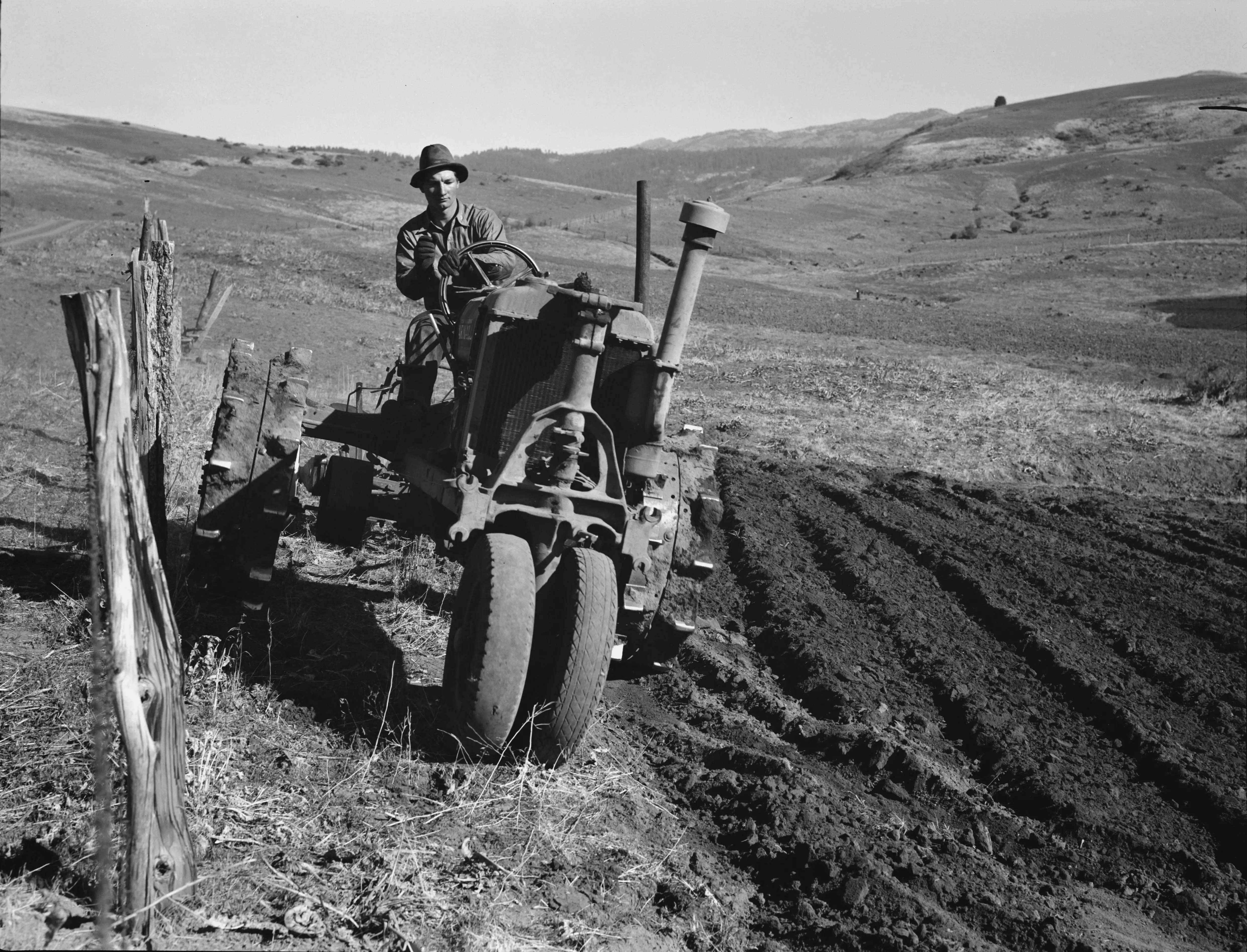 Young farmer plowing while other co-op members work in the sawmill. The tractor does work for five member families. Ola self-help sawmill co-op. Gem County, Idaho.