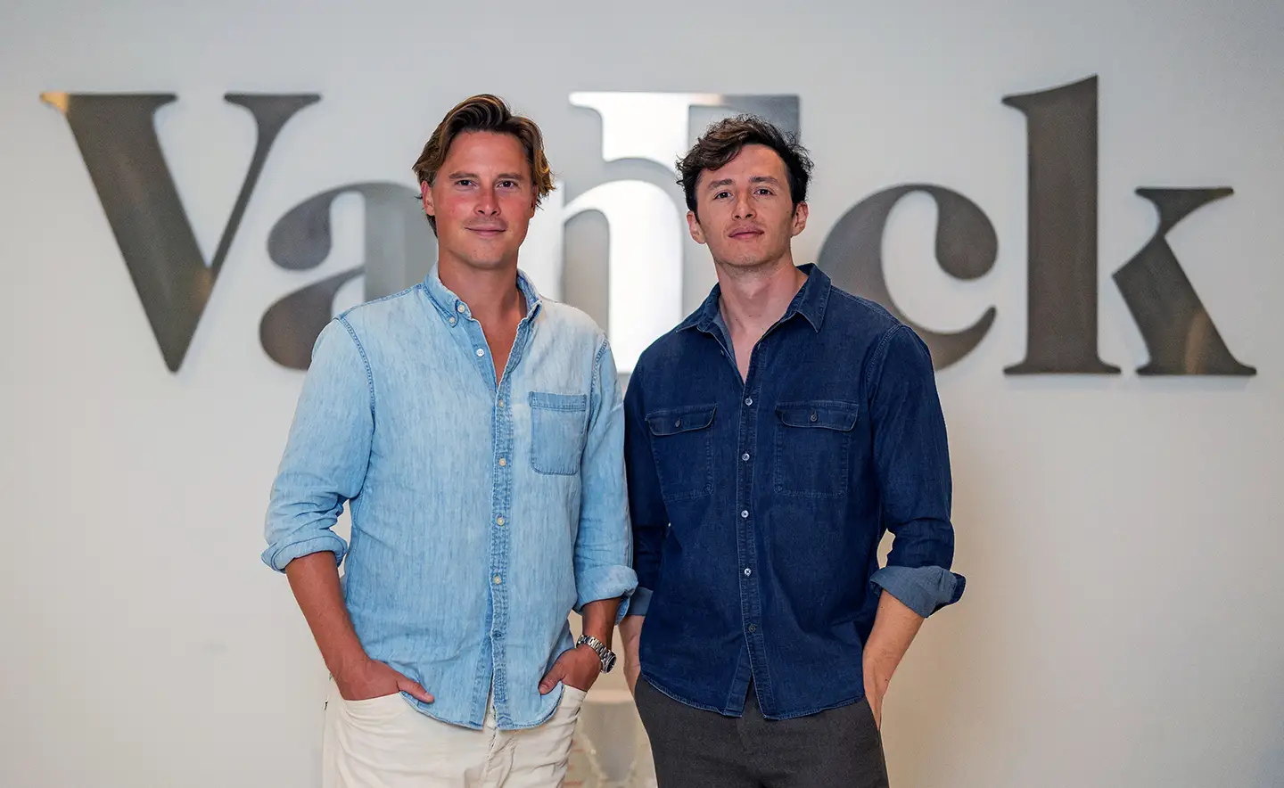 Two men in denim shirts pose in front of a corporate VanEck office sign