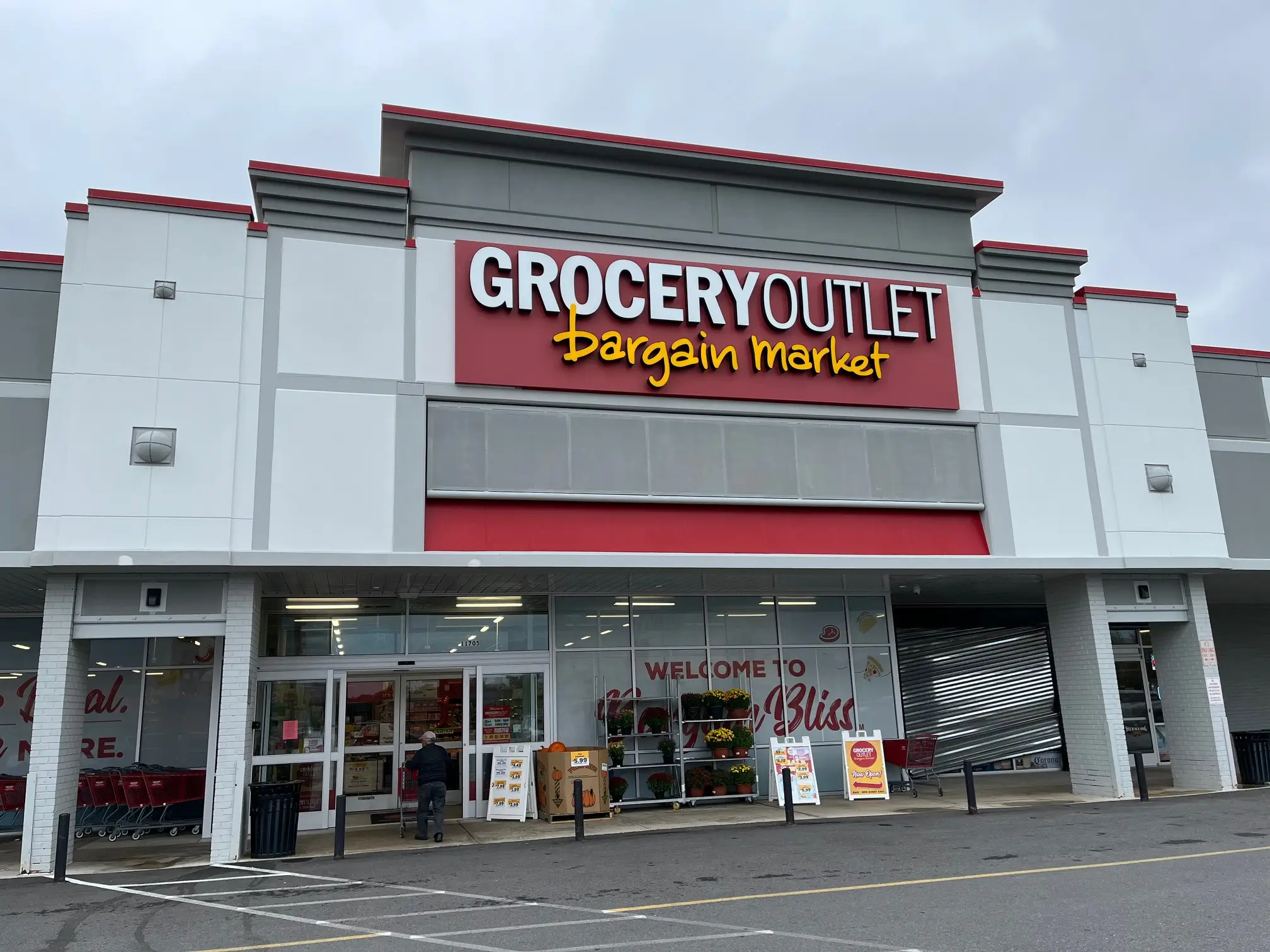 The entrance to a Grocery Outlet store, with a customer pushing a shopping cart into the front door and flowers on a shelf outside.