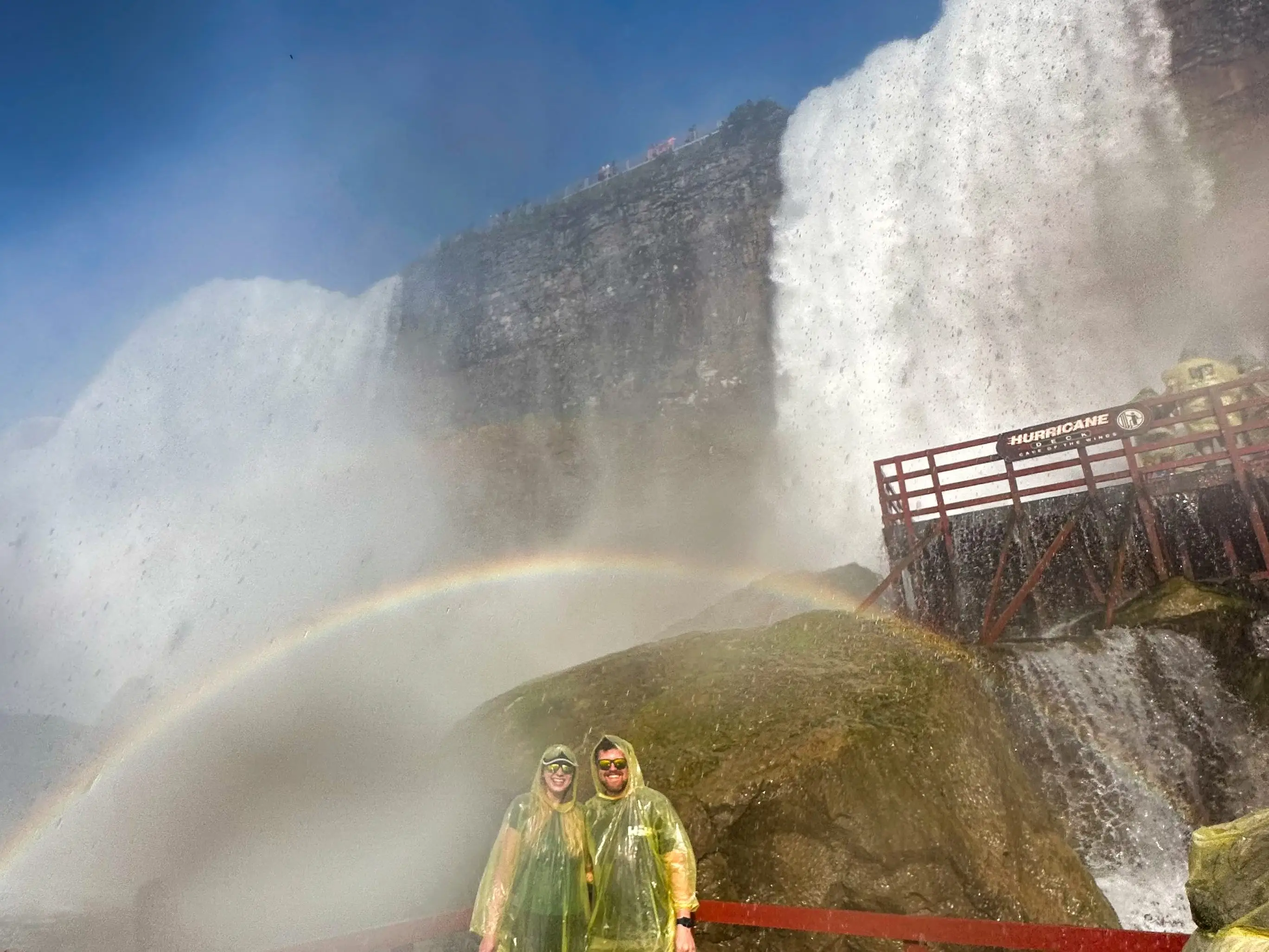 Emily and her husband wear yellow ponchos and stand under a rainbow generated by a waterfall.