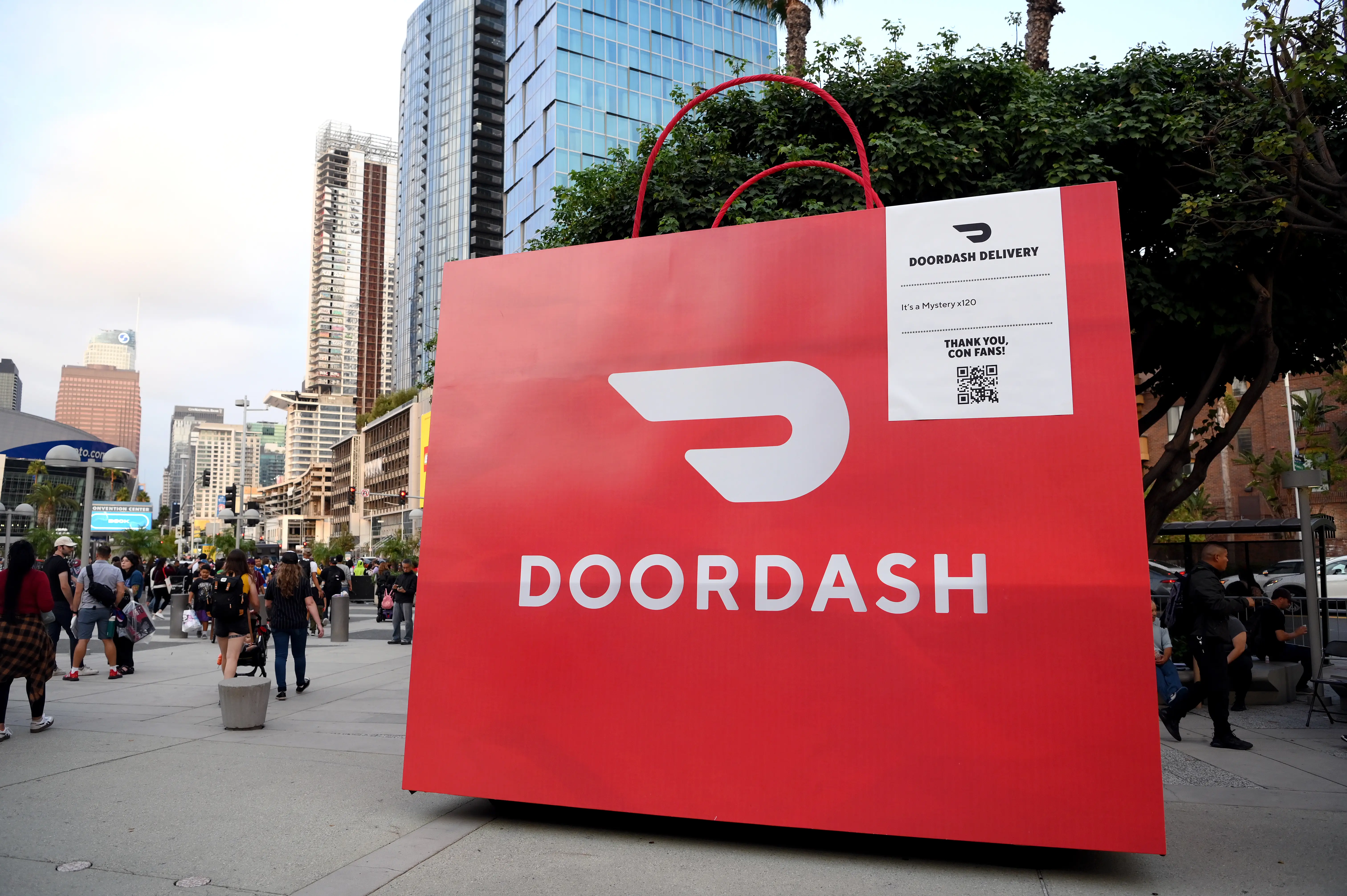 A large sculpture of a red DoorDash delivery bag sits on a sidewalk in Los Angeles as people walk by.