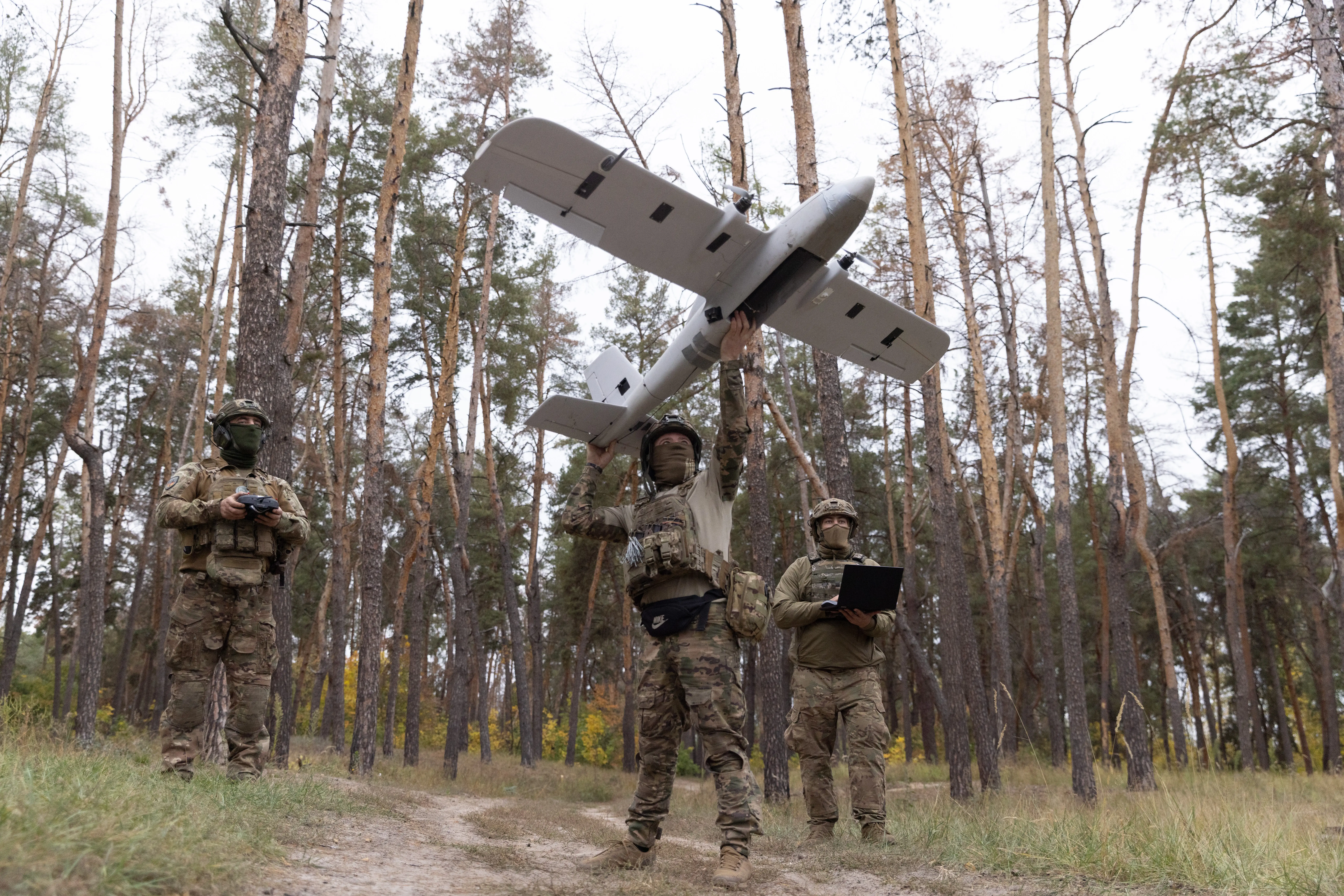 A man in camouflage gear holds a large grey drone above him while surrounded by trees as two other figures stand beside him