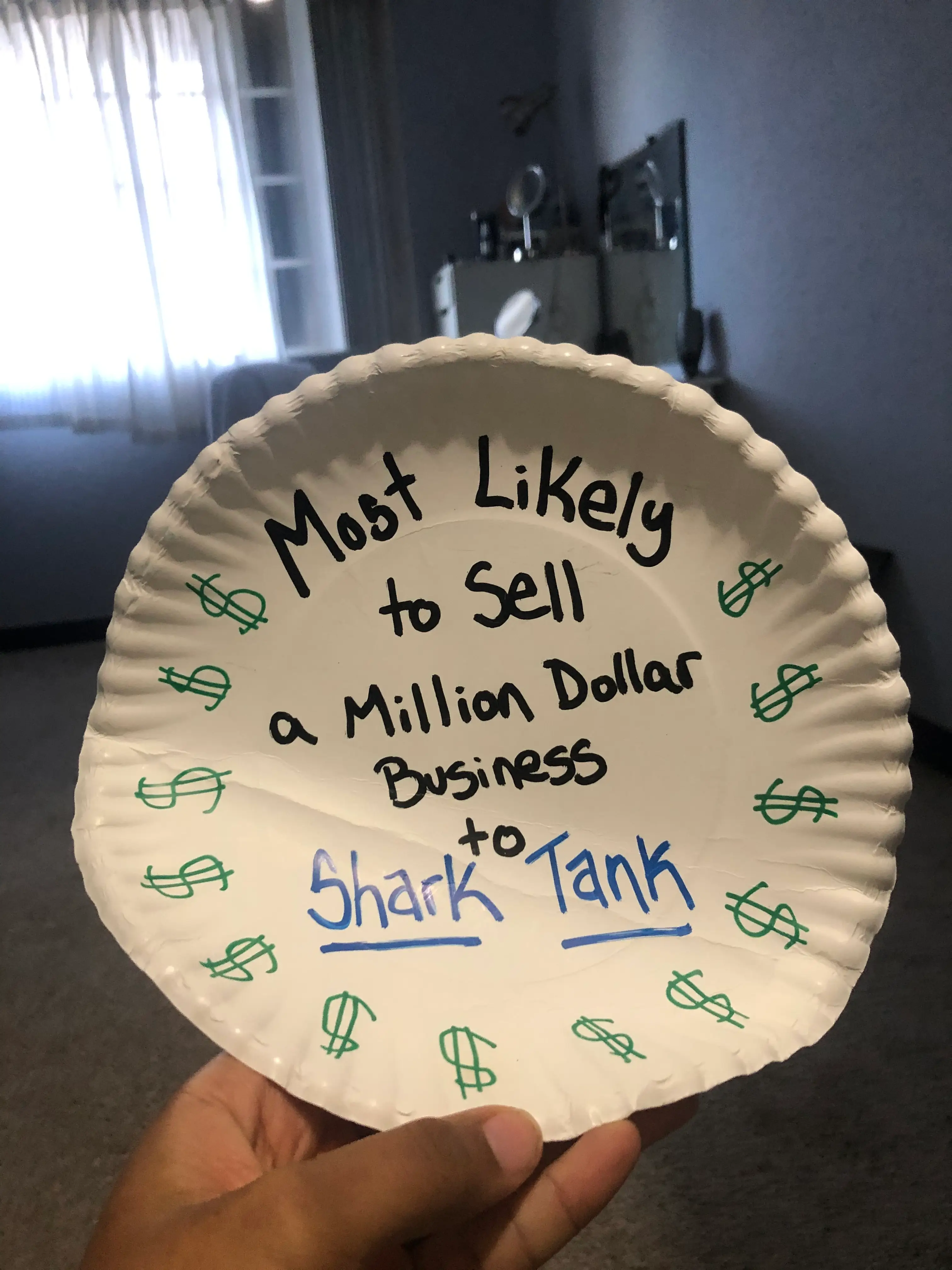 A hand holds a paper plate with writing on it, which reads