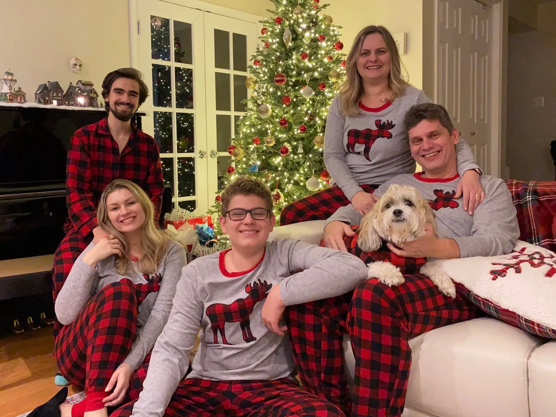 A family in matching Christmas pajamas sits together in front of Christmas tree.