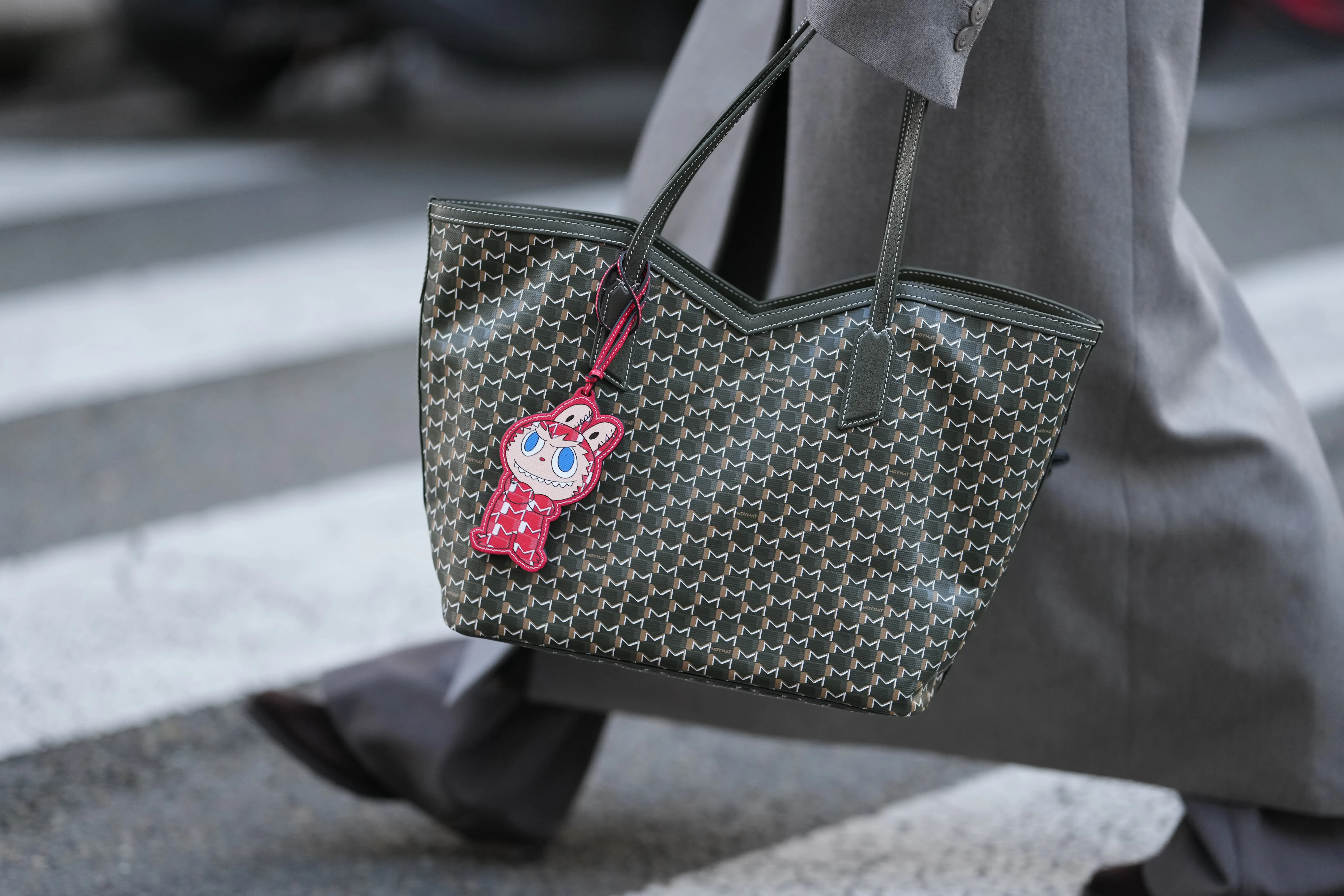 Close-up detail view of a green monogram Moynat tote bag in leather with a red Labubu x Moynat keychain charm, during a street style fashion photo session, on October 27, 2025 in Paris, France.