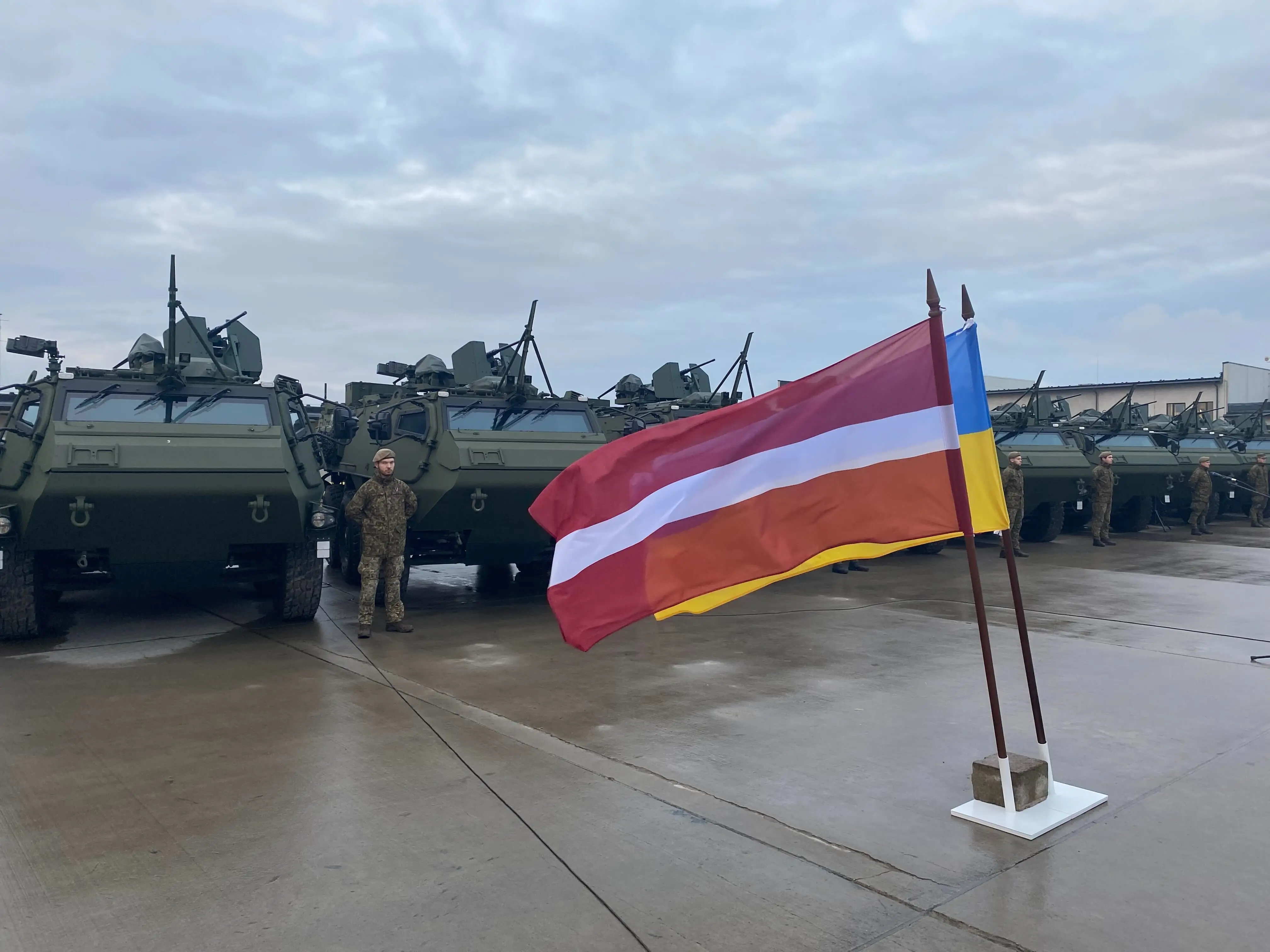 Soldiers stand to attention next to the Latvia and Ukrainian flags, placed before a row of 6x6s.