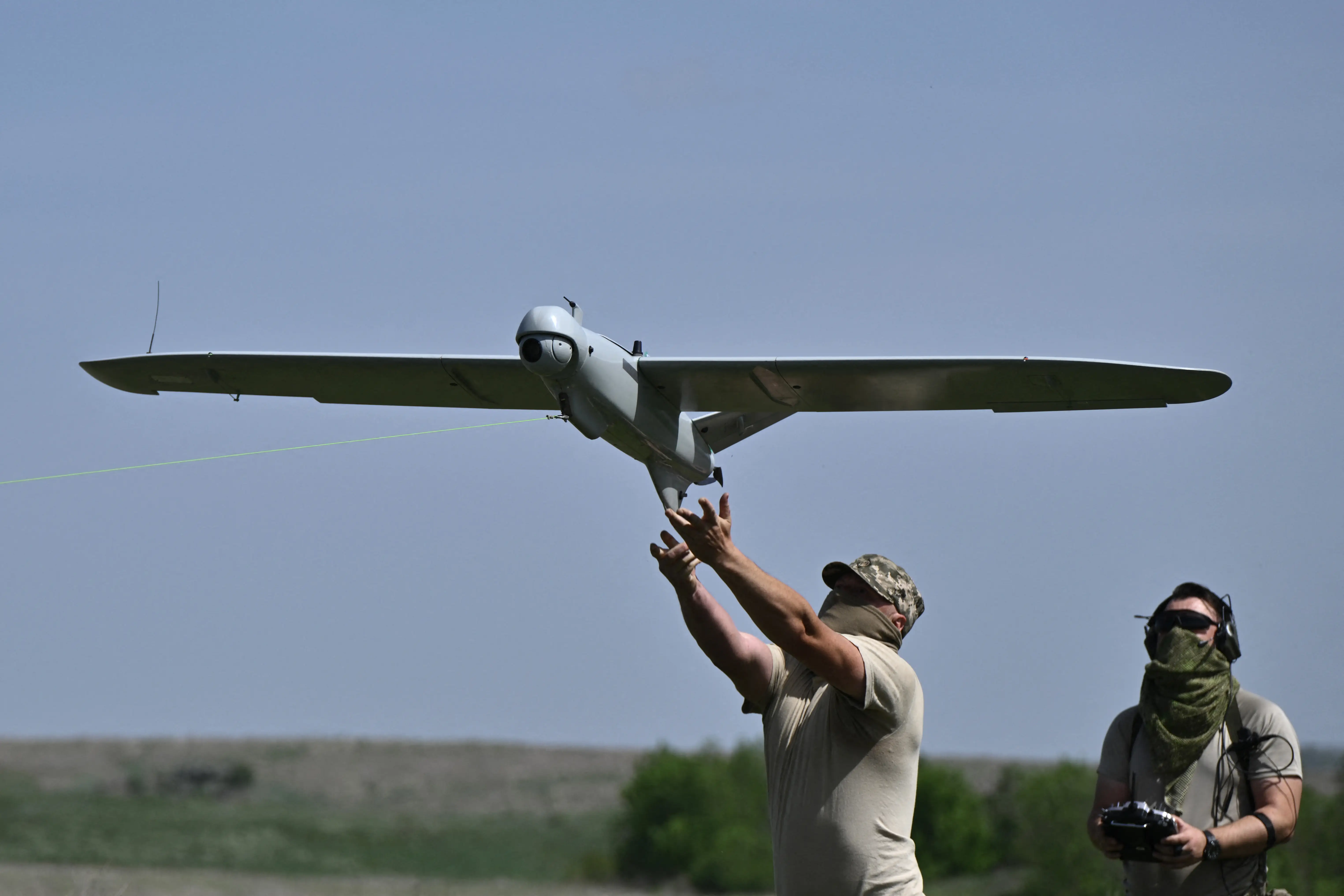A man in grey clothing launches a large grey drone into the sky with another man holding a controller behind