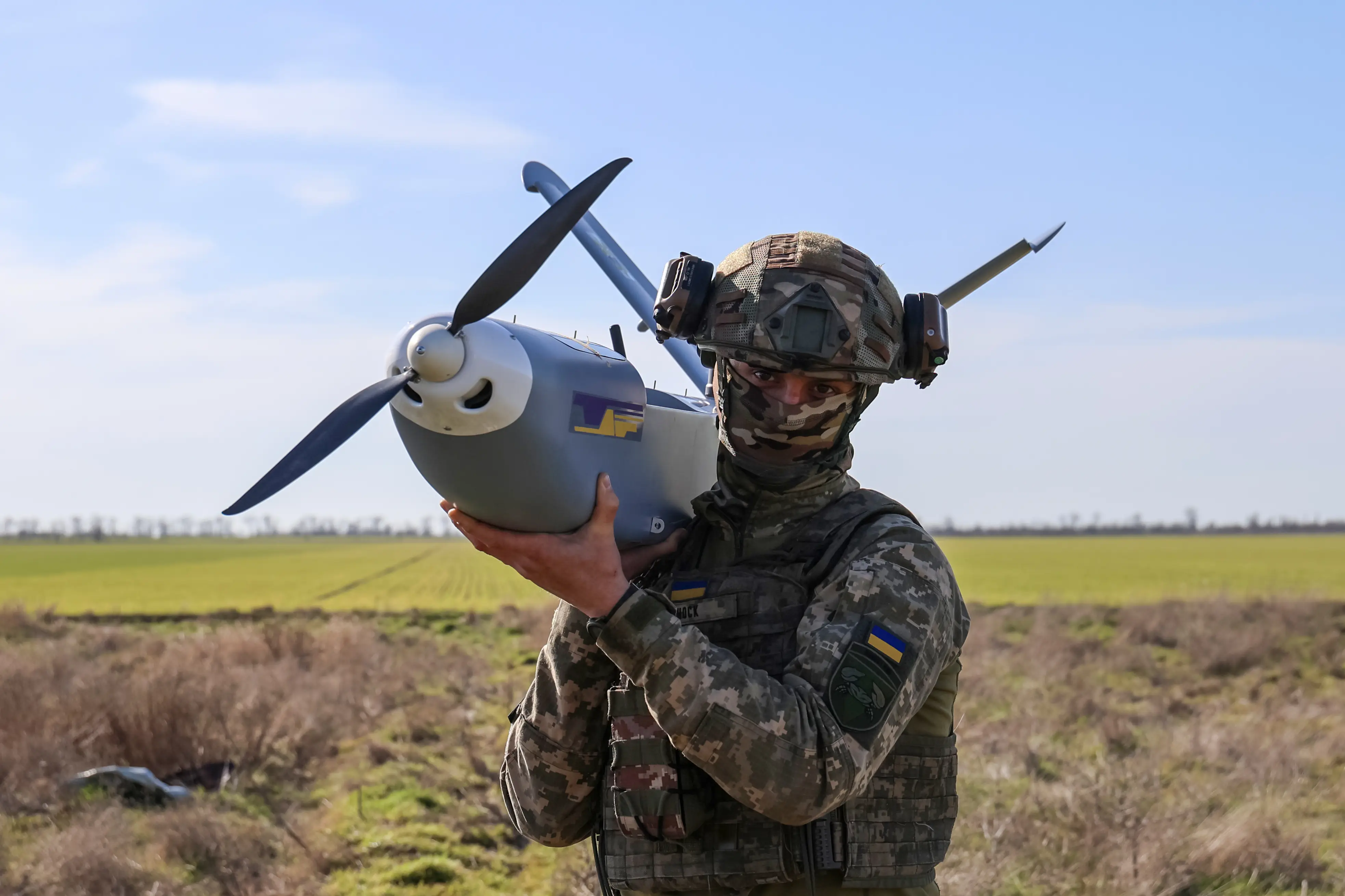 A man in camouflage gear holds a large grey drone while looking at the camera in a sunny field