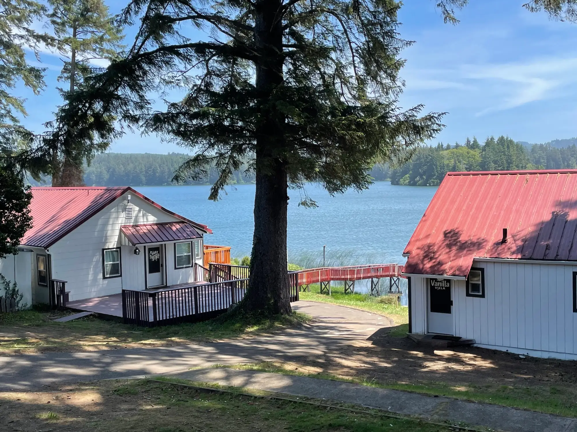 Two white cabins with red roofs. Each has a patio, and a red bridge leads to a body of water.