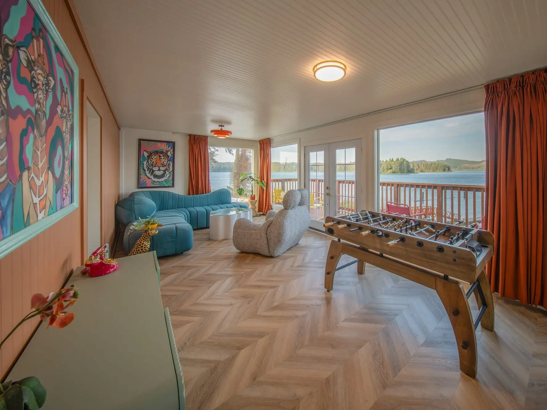 A living area with a large patio that overlooks a lake. A blue, curved couch, a char, and a foosball table sit in the space.