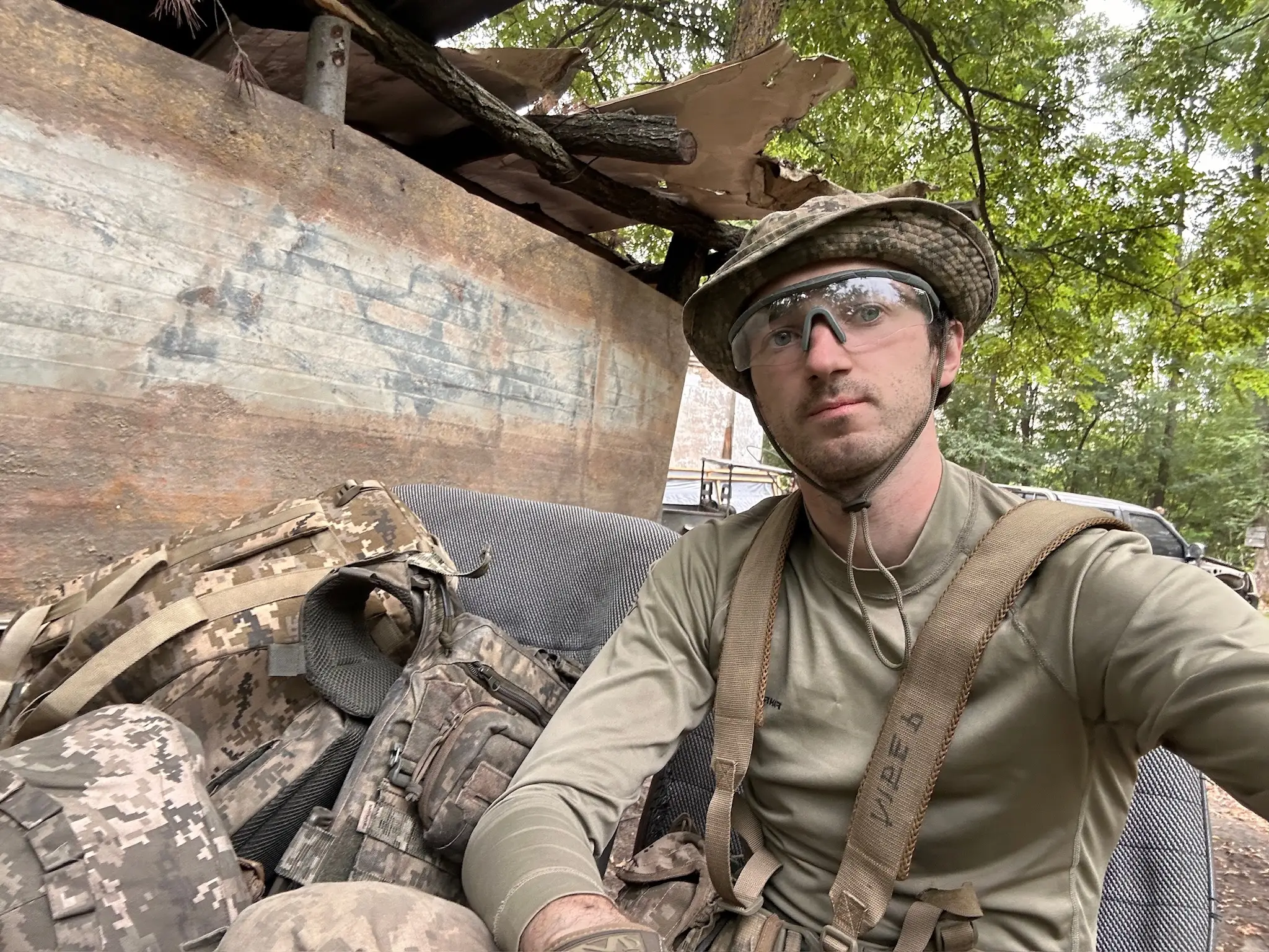 Dimko Zhluktenko, with a jungle hat and protective glasses, is seen sitting with his gear in a forest.