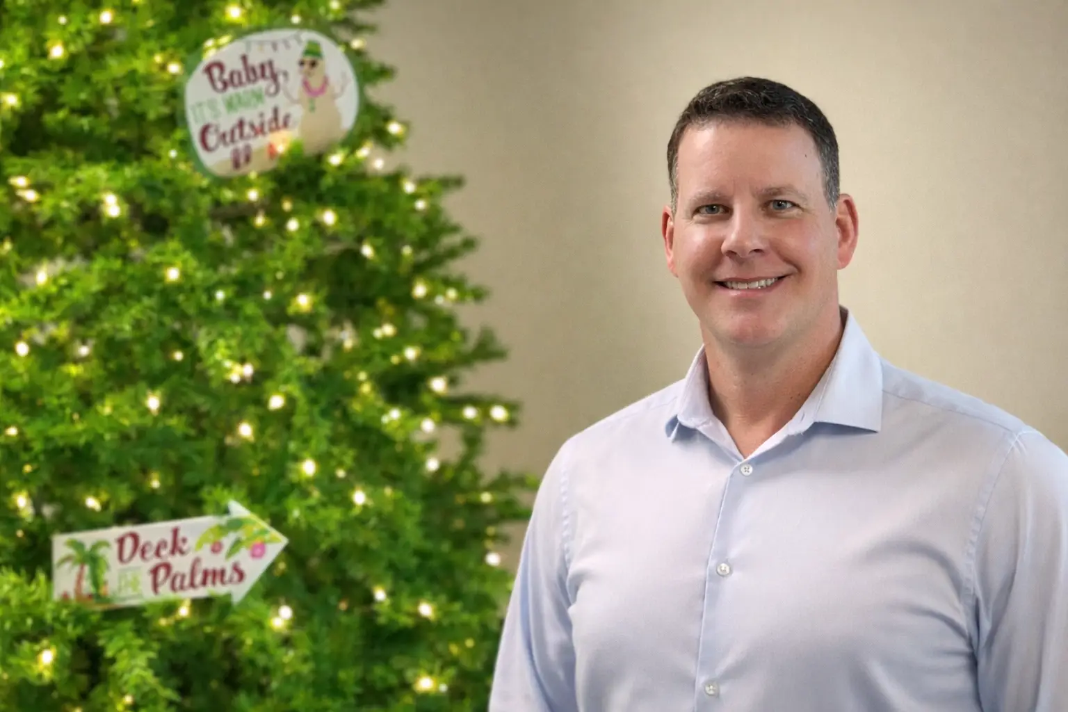 A man in a button-down shirt smiles beside a Christmas tree