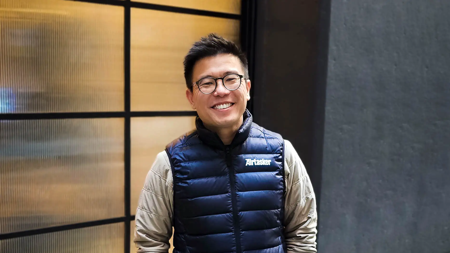 Tim Fung, founder and CEO of Airtasker, smiles while wearing a dark blue puffy vest and a light grey shirt and glasses with black rims.