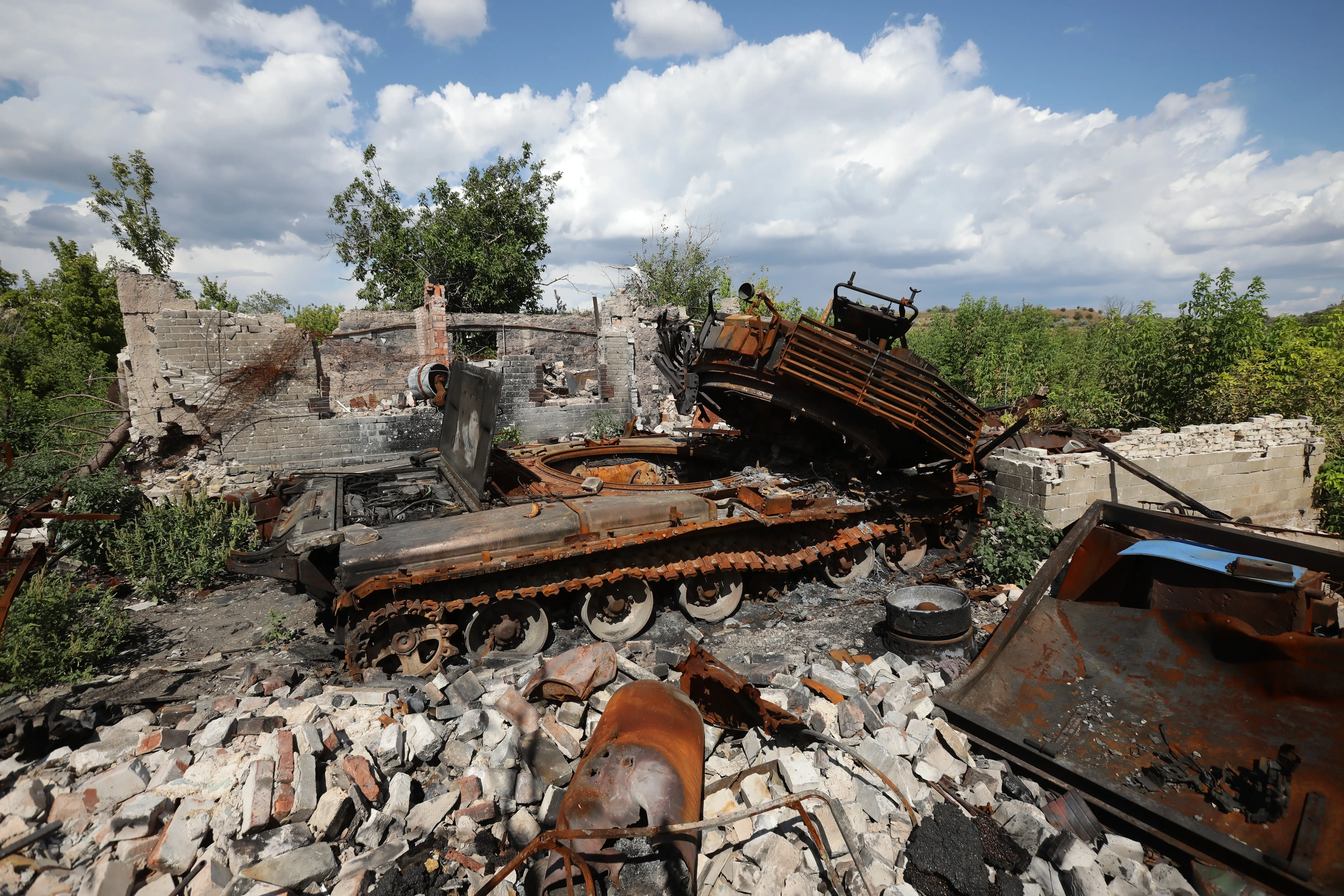 A rusted destroyed tank on top of bricks and beside a destroyed house under a blue sky