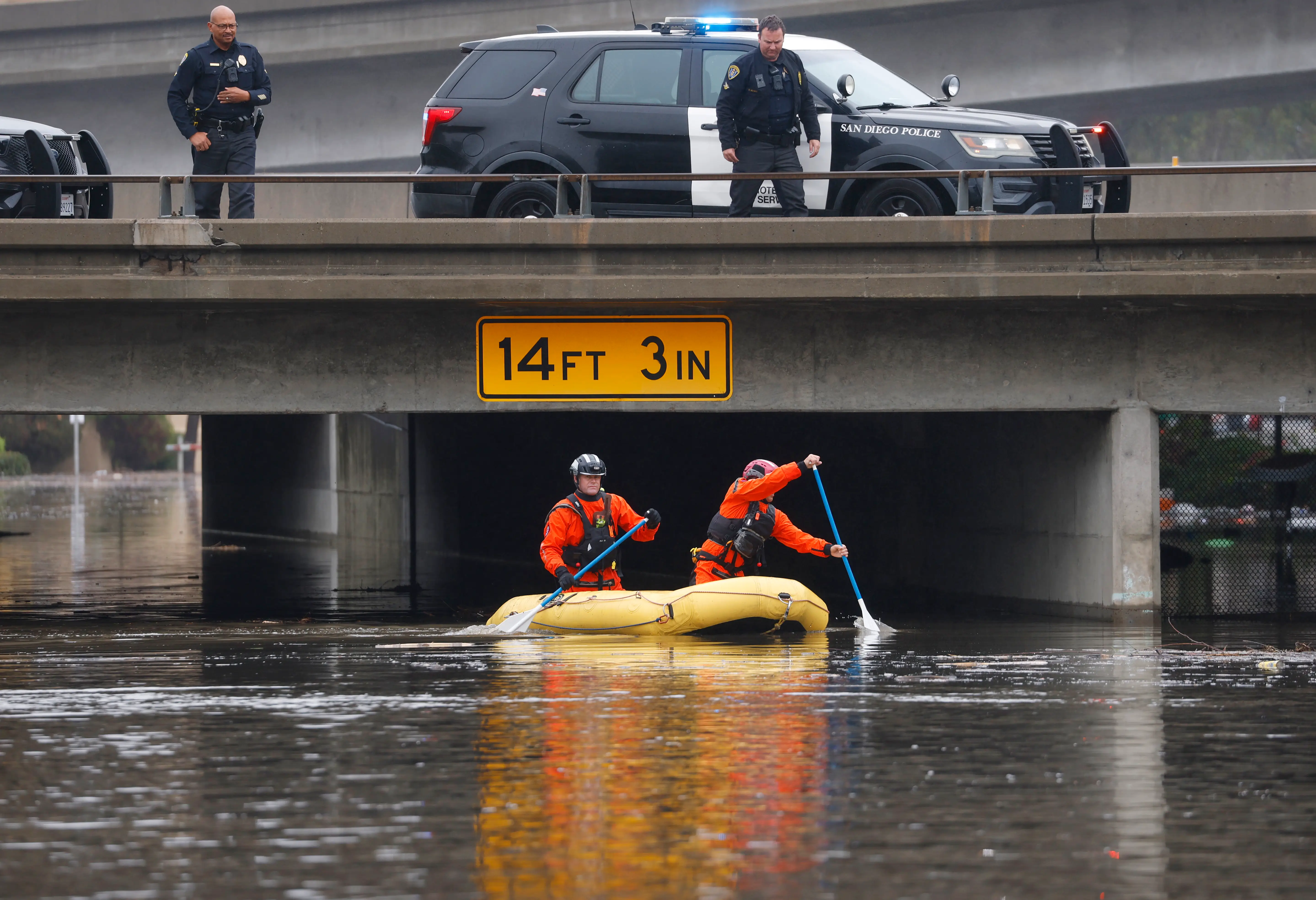 : San Diego Police officers, standing on State Route 163, and members of the San Diego Lifeguard River Rescue Team look for people after the San Diego River overflowed and flooded areas in Mission Valley during a storm on January 1, 2026 in San Diego, CA.