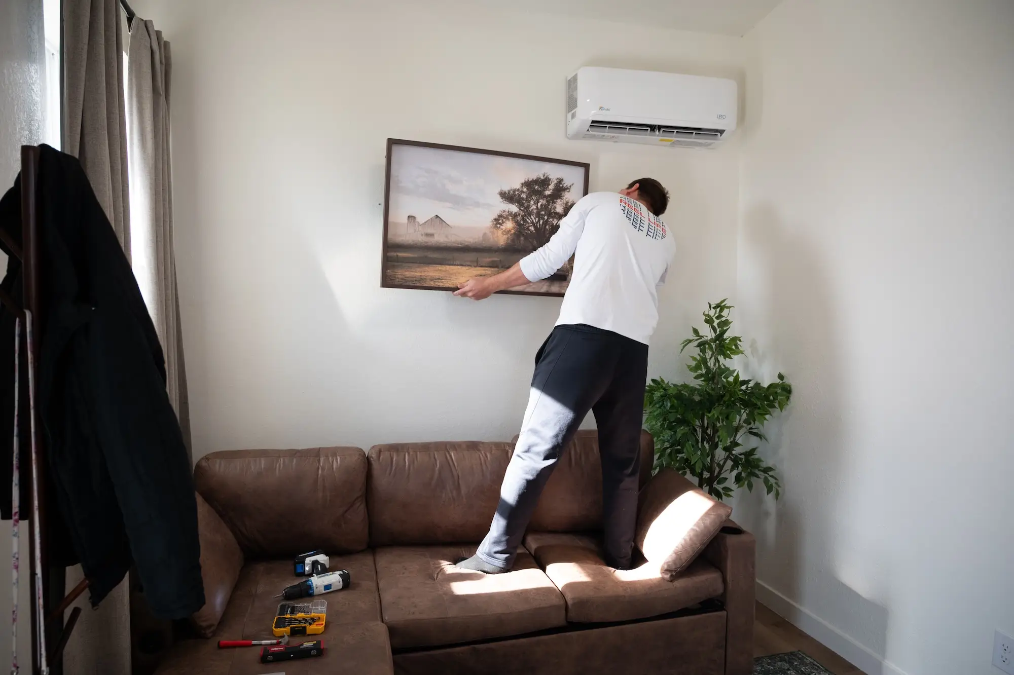 A man stands on a couch to hang a painting above a couch.