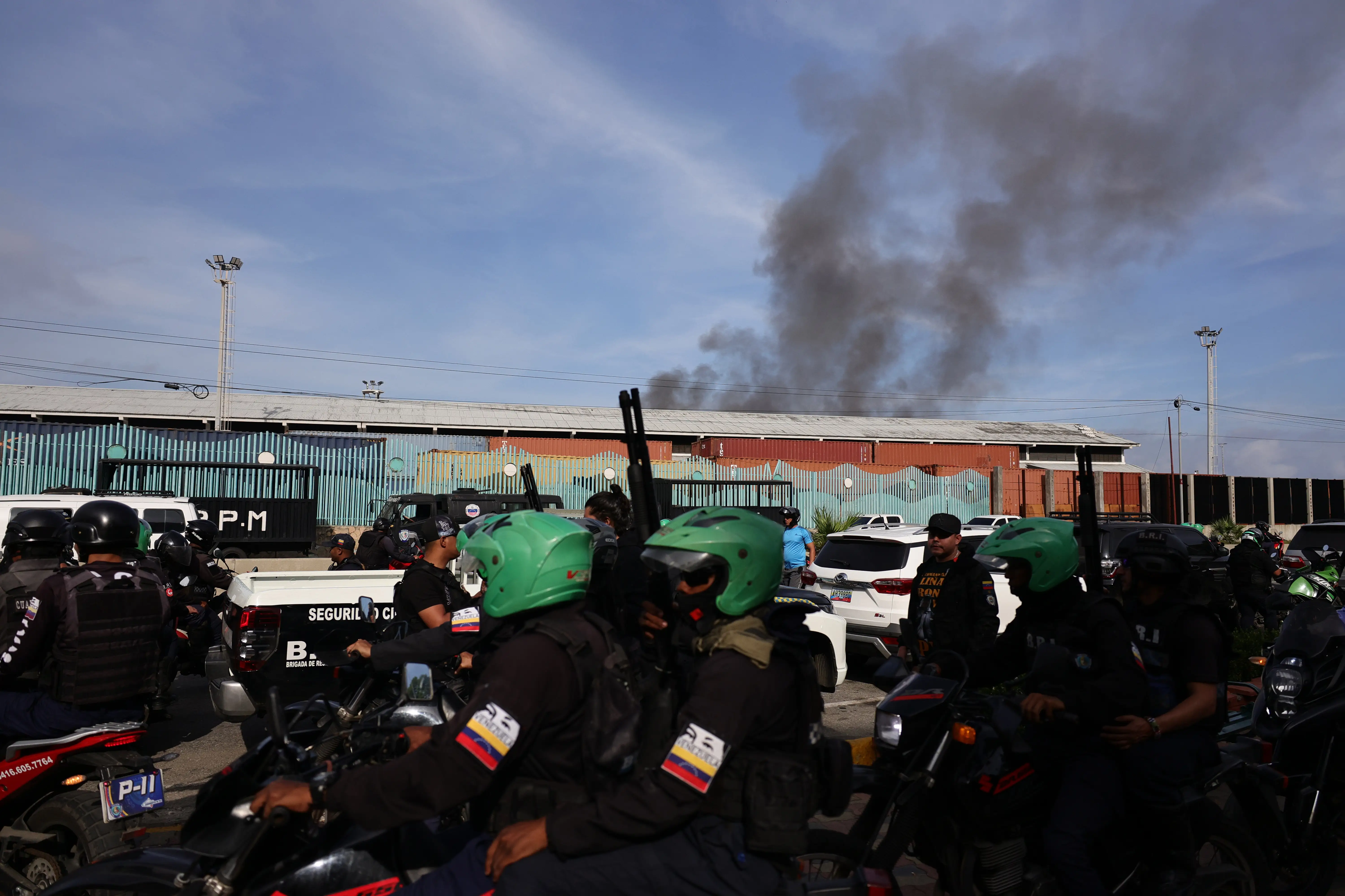 Groups of people ride motorcycles and hold guys outside of a port in Venezuela. There is smoke rising in the background from a strike.