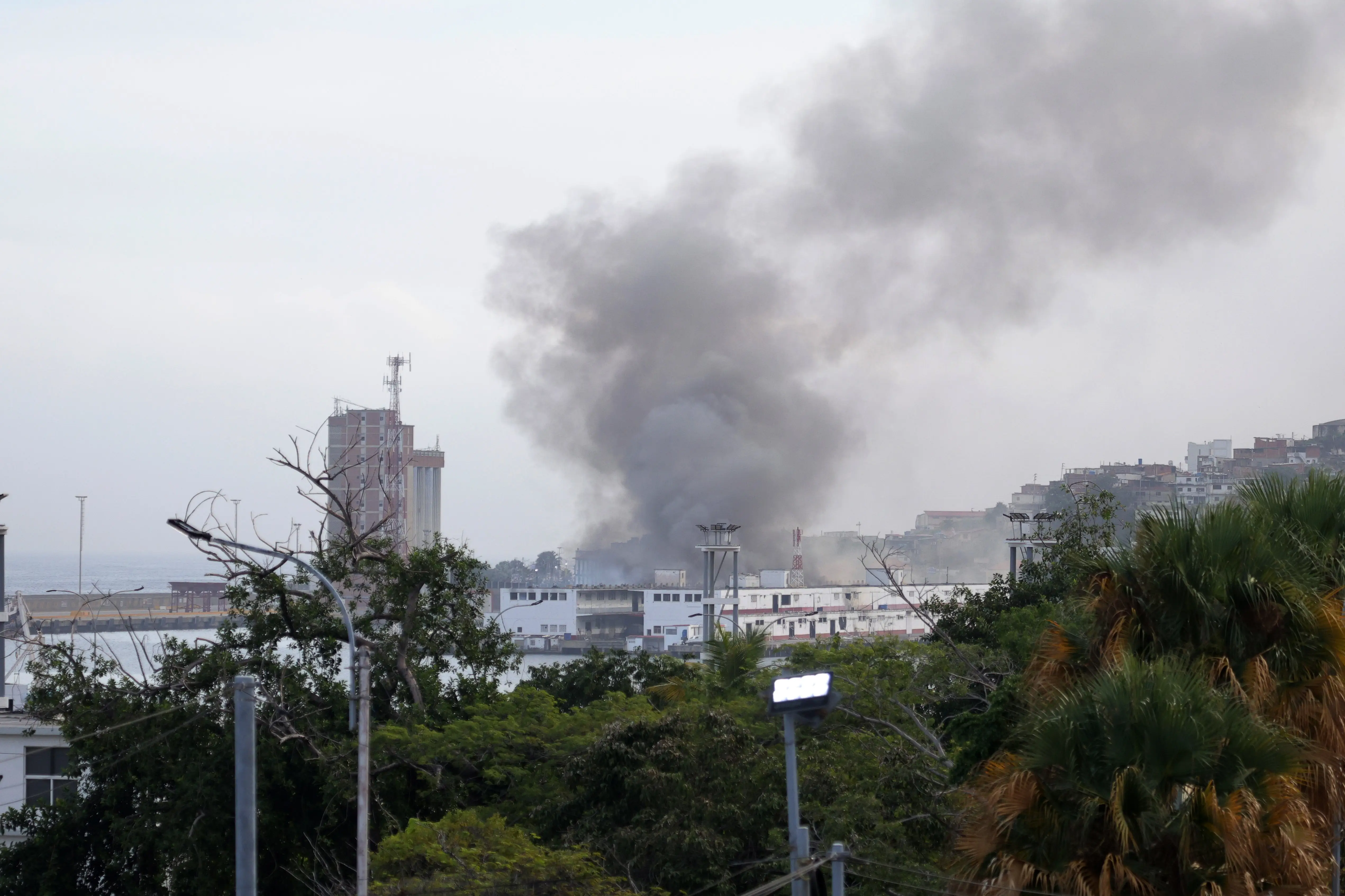 Grey smoke rises from a white building. Other buildings are seen in the background. In the foreground there is a line of green trees.