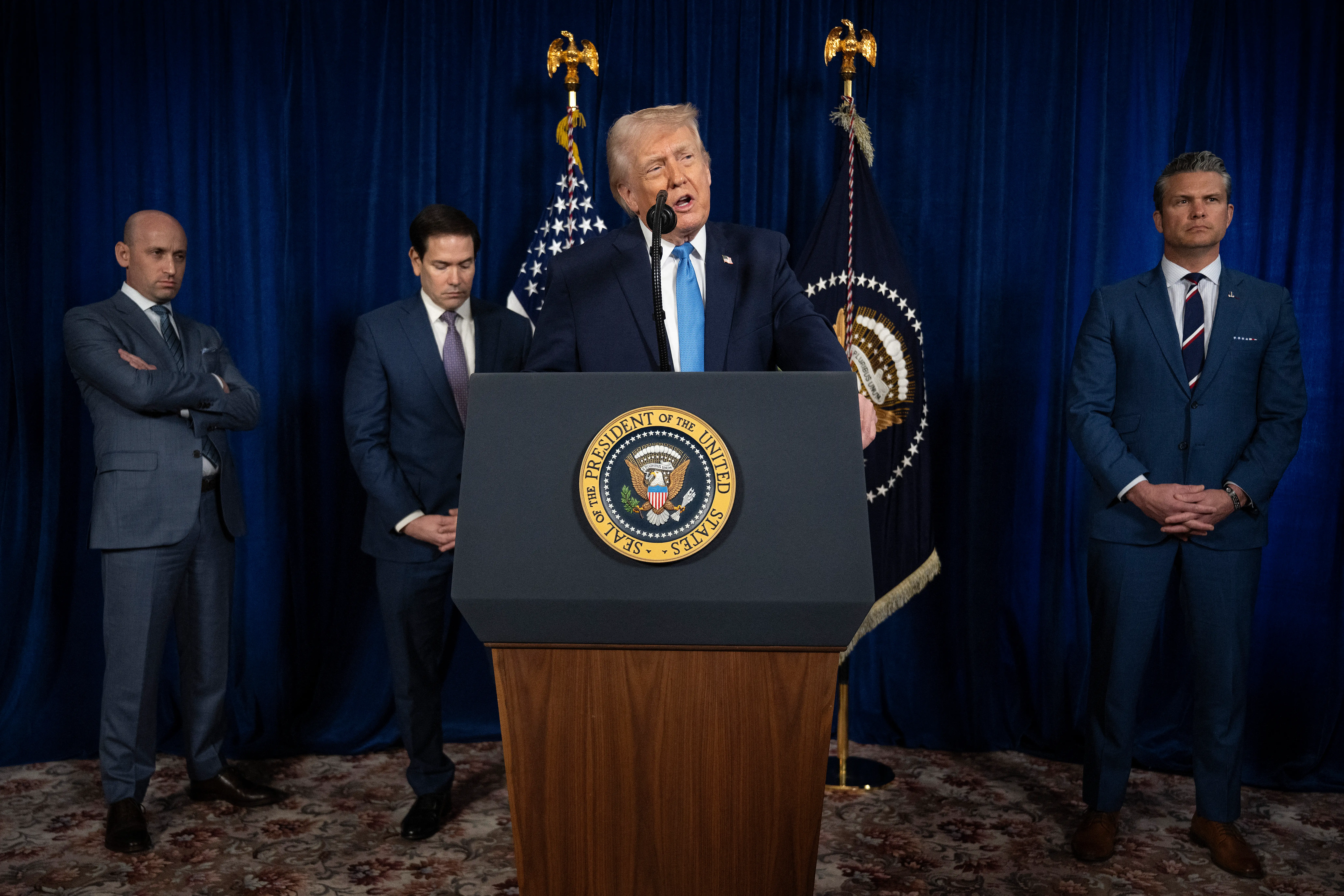 President Donald Trump speaks at a podium about his January 3rd raid on Venezuela. Pete Hegseth, Marco Rubio, and Stephen Miller stand behind him.