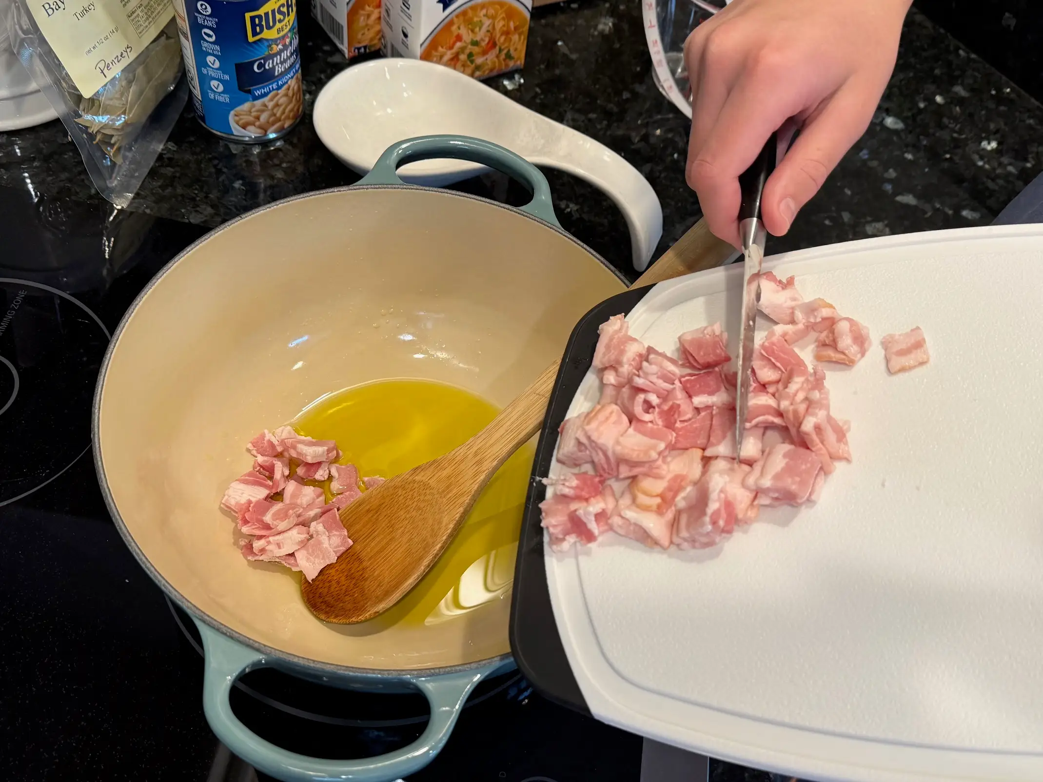 Kristina uses a cutting board and a knife to add diced bacon to a pot with olive oil.