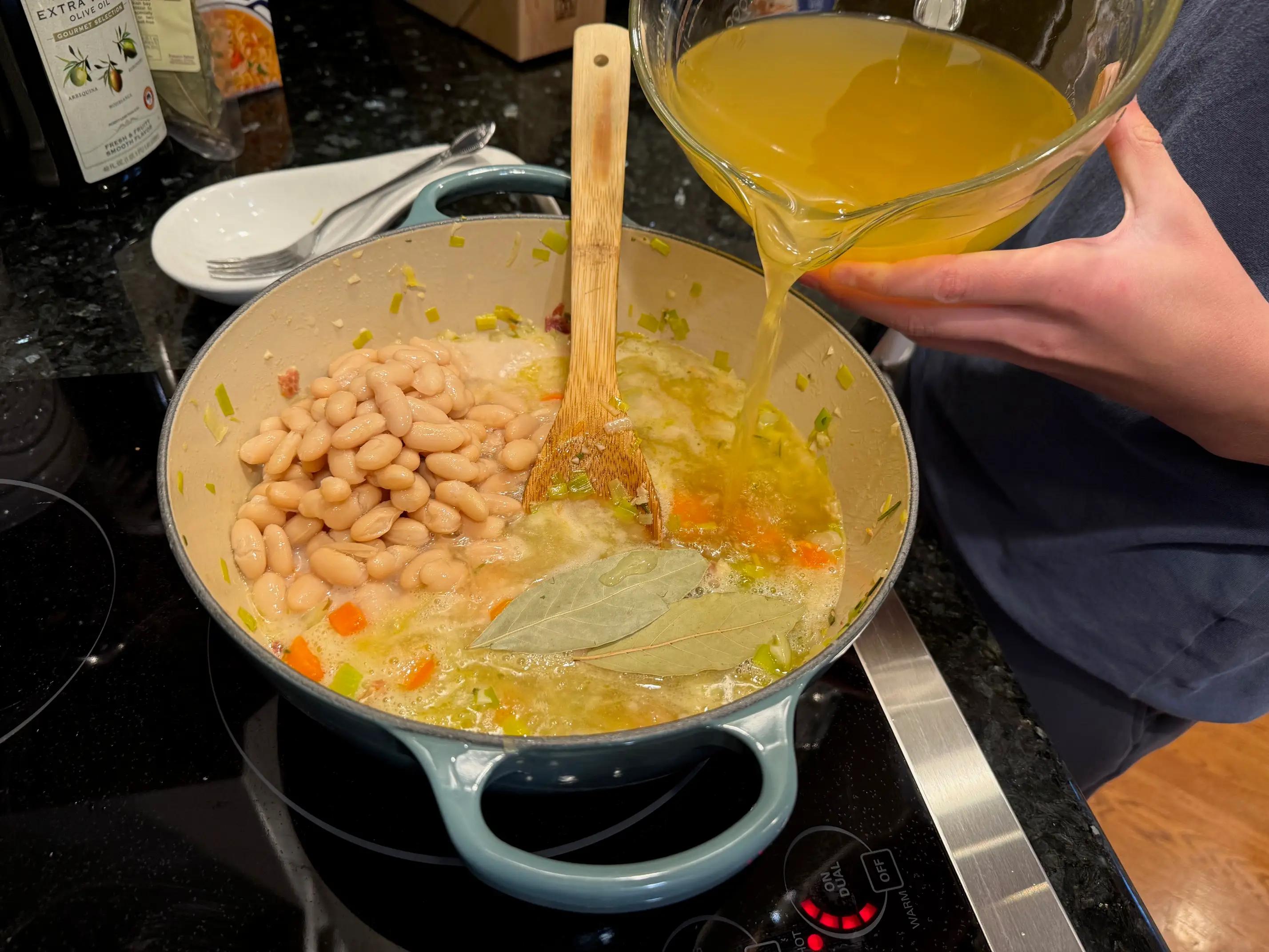 Kristina pours broth into a pot with the vegetables, whole and pureed cannellini beans, and bay leaves.