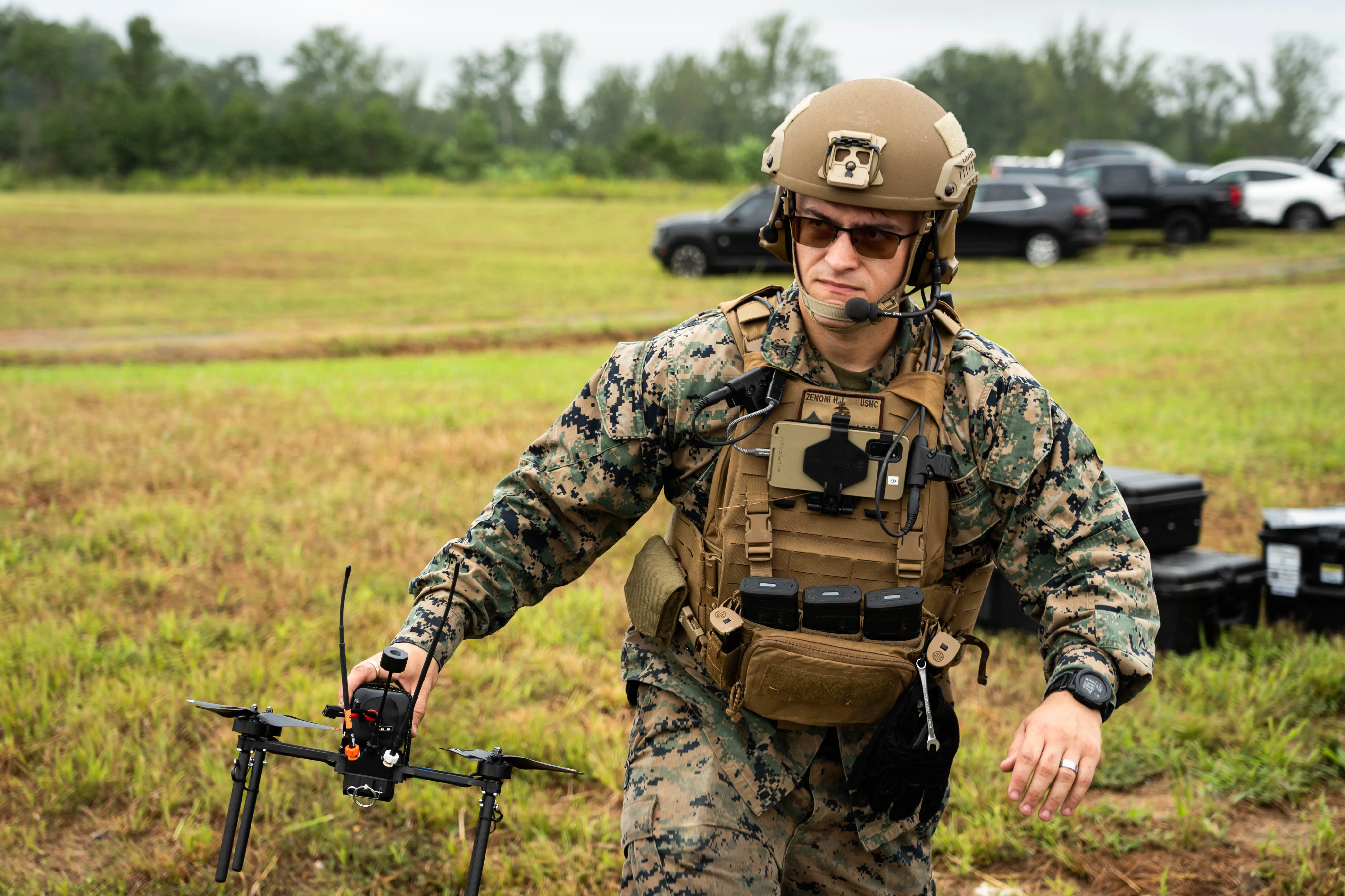 A man wearing sunglasses and a camouflage uniform carries a small black drone.