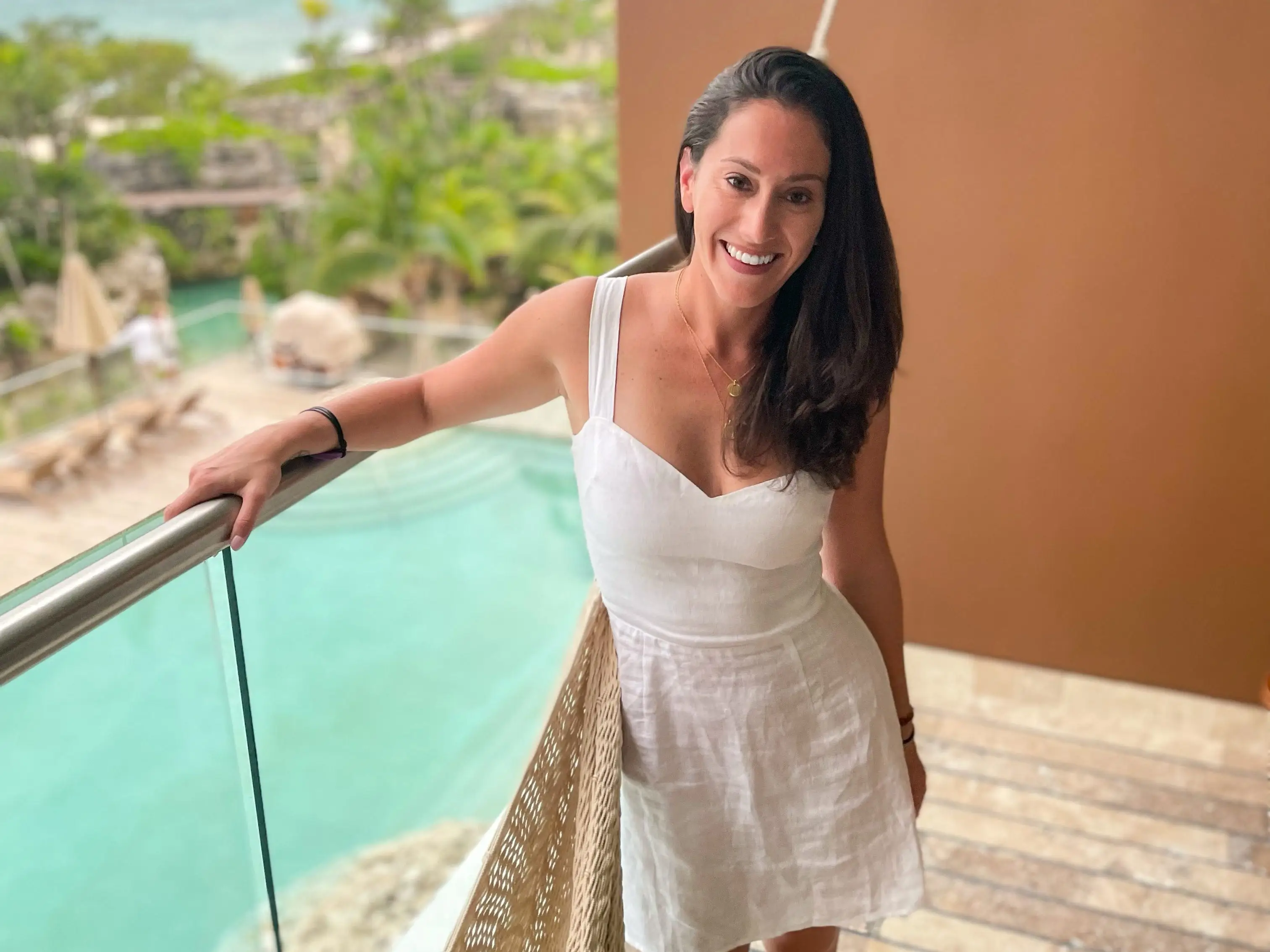 A smiling woman in a white dress leans on a balcony railing overlooking turquoise water, palm trees, and a seaside resort below.