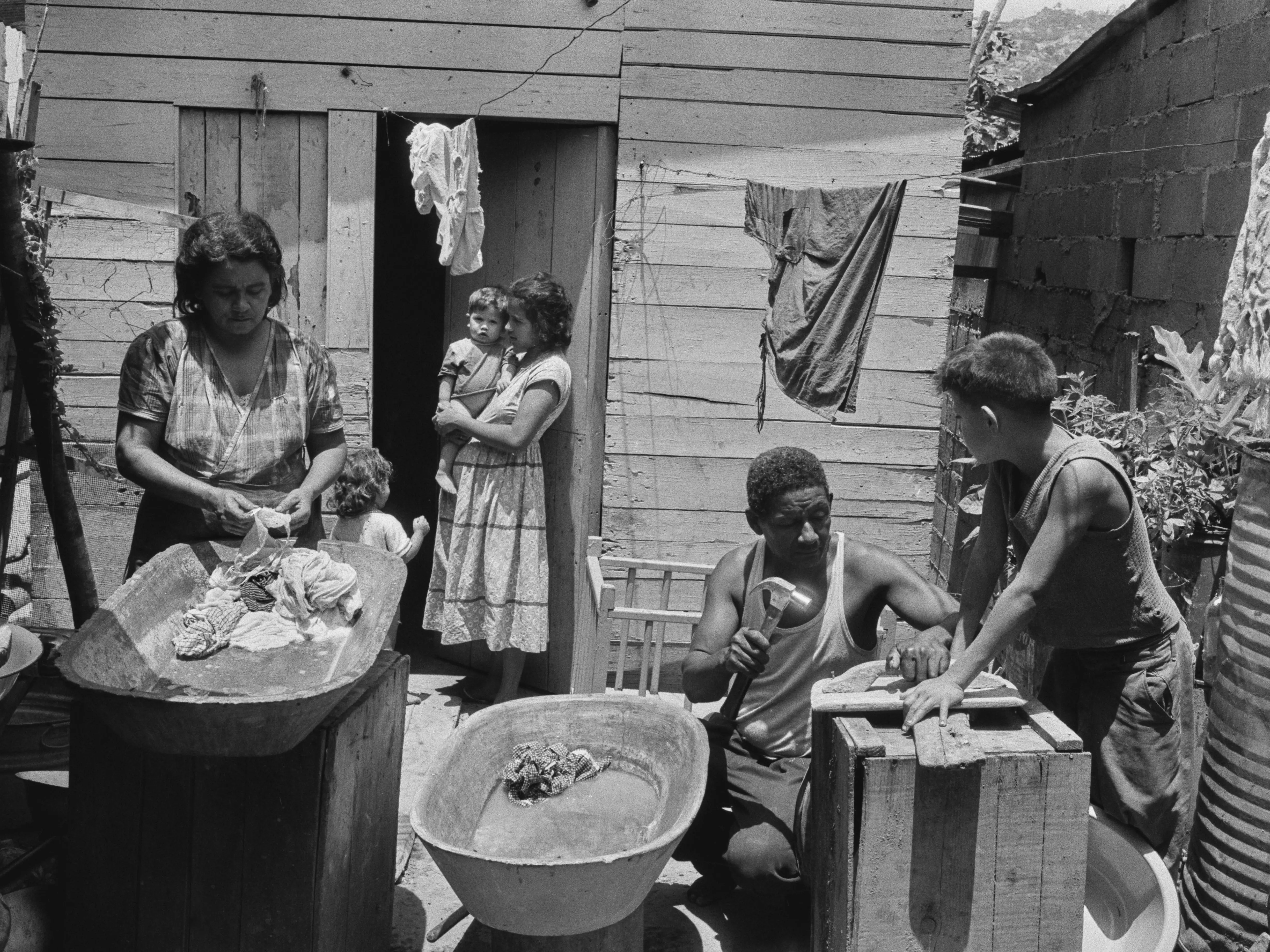 A woman doing the laundry in a washtub as a young woman stands in a doorway holding a young child, and another child assists a man with repairs outside a wood-built dwelling in Caracas, Venezuela, circa 1957.