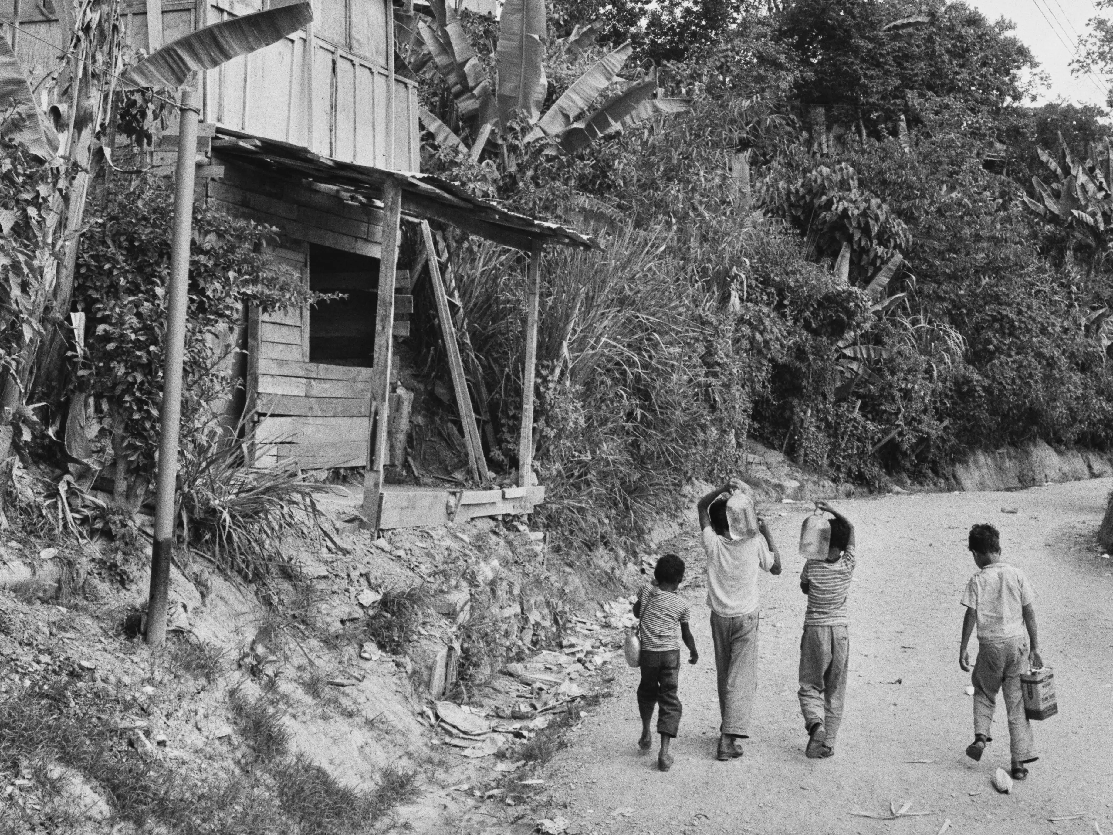 A group of children passing a wood-built structure as they carry bottles and an oil tin in the village of Petare on the outskirts of Caracas, Venezuela, circa 1957.
