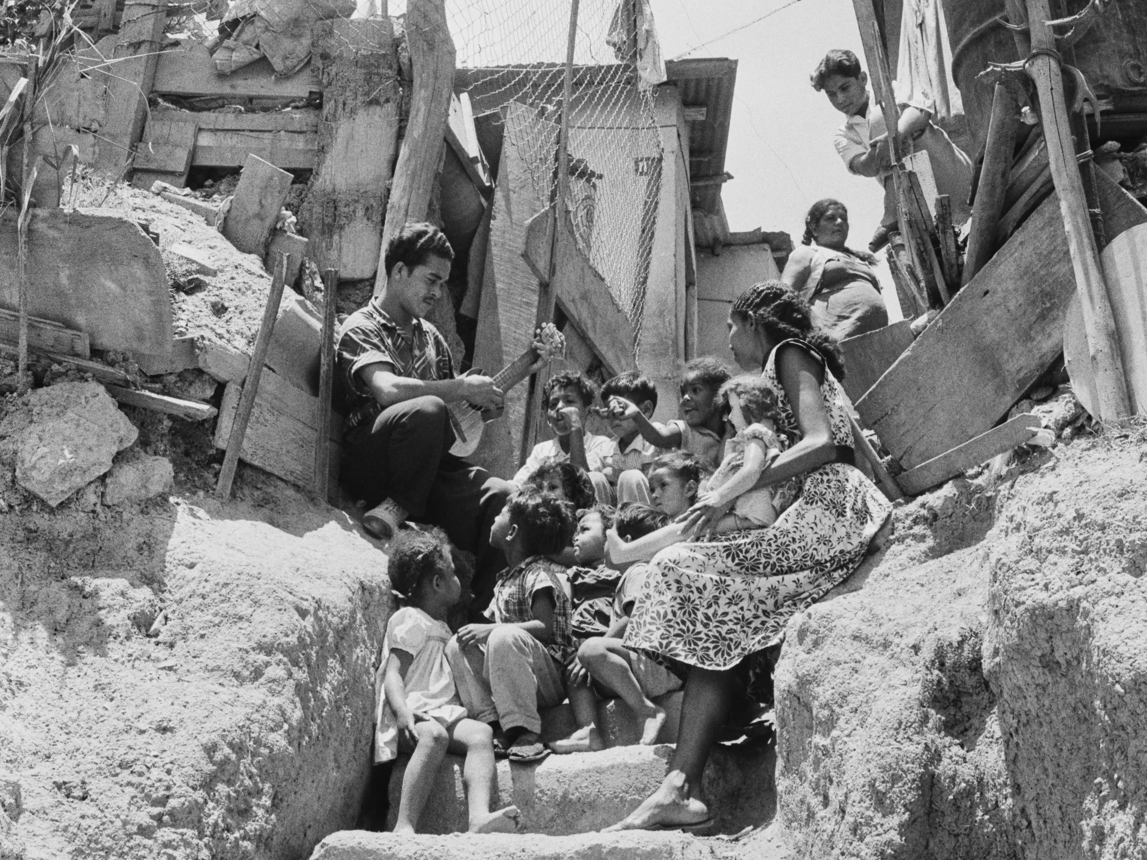 A man plays a cuatro for the entertainment of a group of children and a woman, all sitting on a flight of steps leading to slum housing, with a washing lines visible through a chain link fence, in Caracas, Venezuela, circa 1957. The Venezuelan cuatro is a four-stringed instrument, similar in shape to the ukulele, played by a cuatrista.