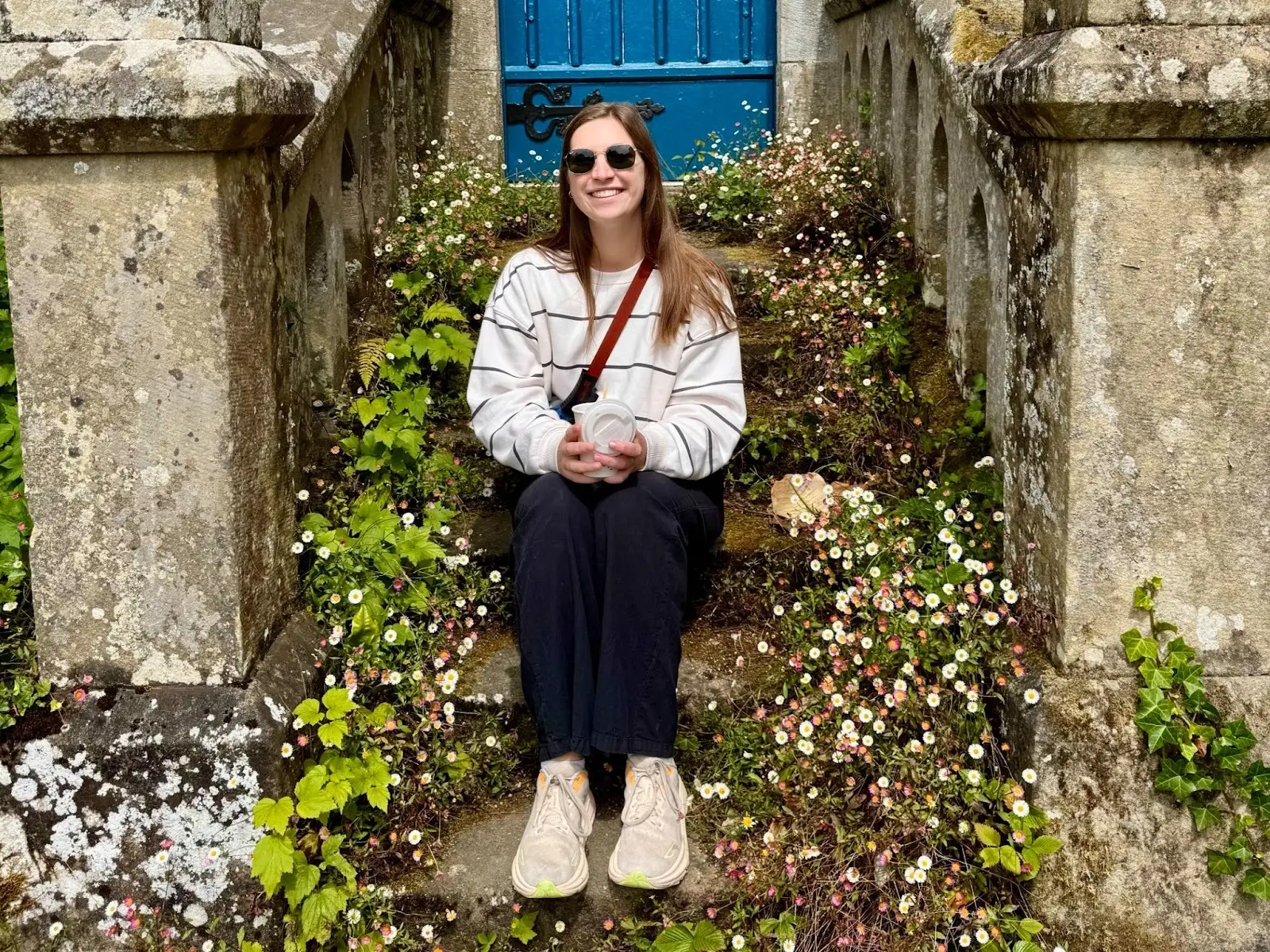 Hannah sits on a stoop covered in flowers, wearing a sweater, black pants, and sneakers.