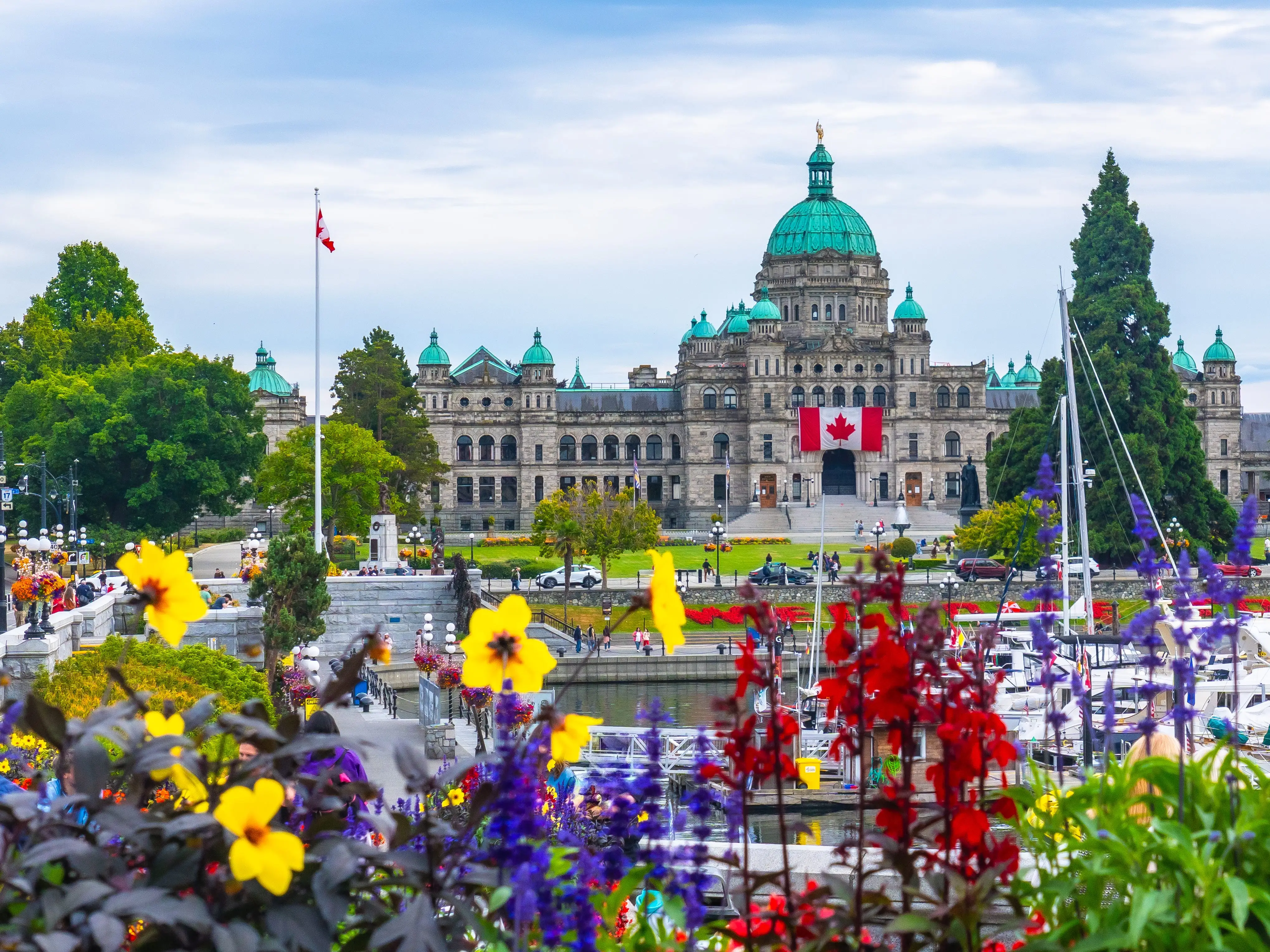 british columbia parliament buildings are seen from across the inner harbour during a beautiful summer day in victoria, british columbia, canada, with colorful flowers in the foreground