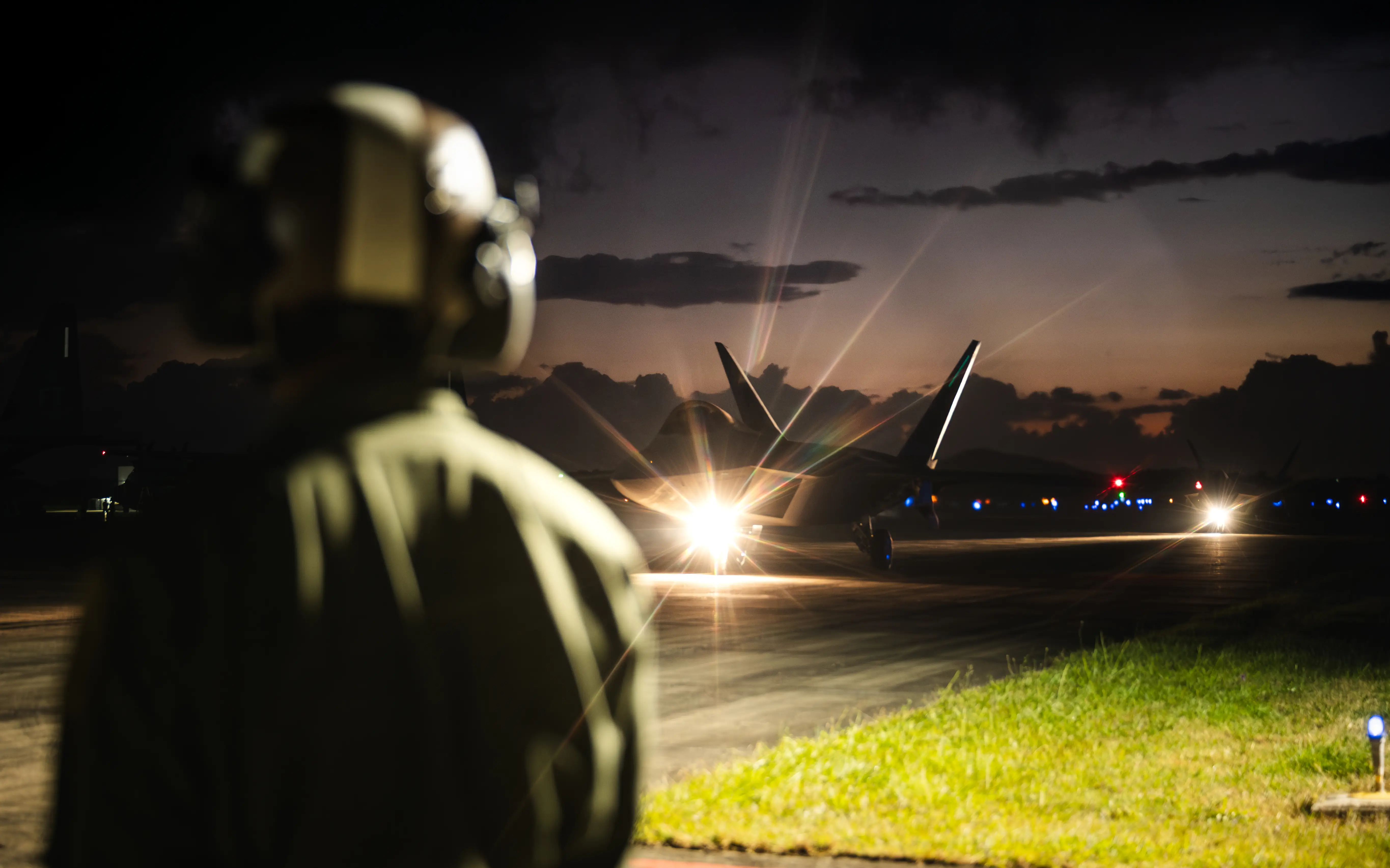 A figure in a helmet stands in front of a fighter jet with lights on at night time