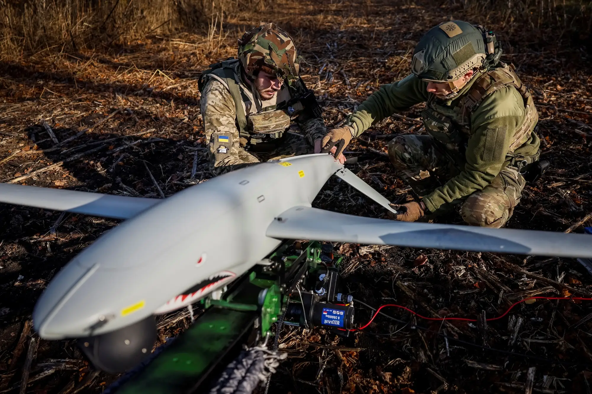 Two men in camouflage gear kneel outside working on a large grey winged drone with a snark's face painted on the front of it