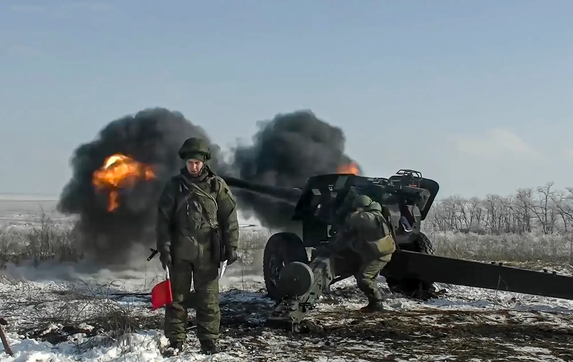 Two figures in green gear stand on snowy ground under a blue sky as a large piece of weaponry fires