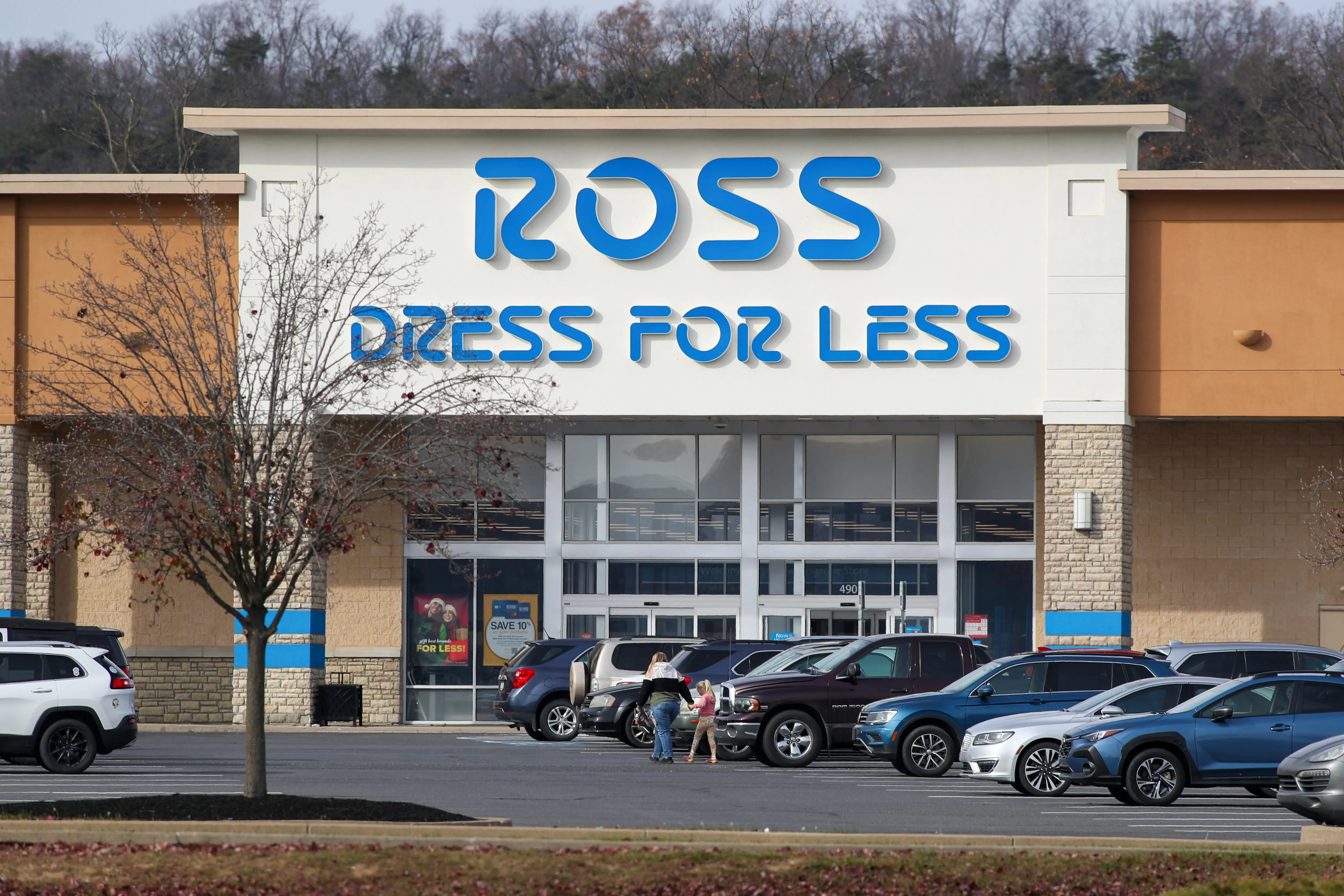 Cars sit parked in front of a Ross Dress For Less store as a woman and child walk through the parking lot and a tree dropping the last of its fall leaves stands in the parking lot.